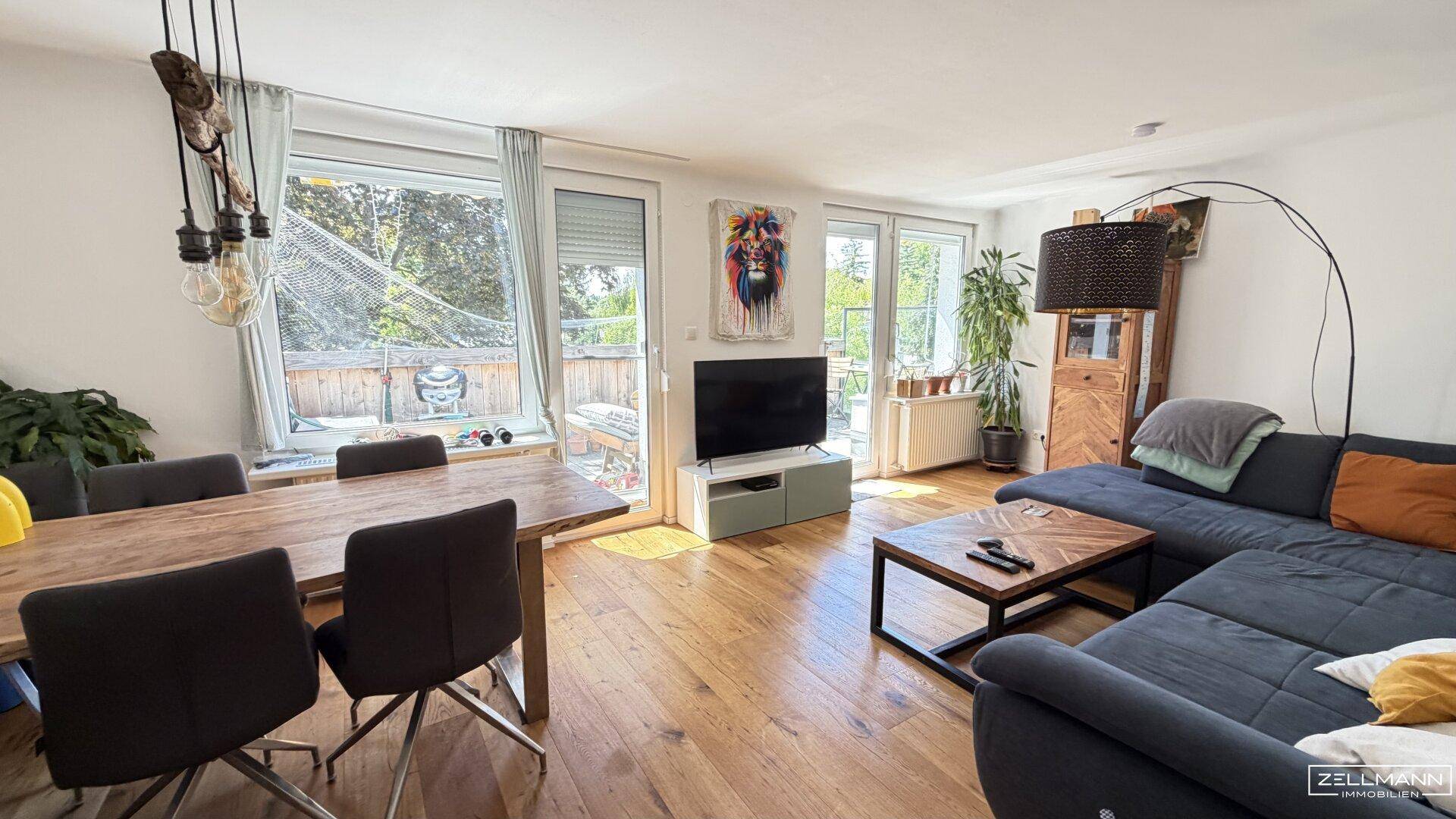 Heller offener Wohn-Essbereich mit Holzboden, großem Sofa und Esstisch mit Balkonzugang.