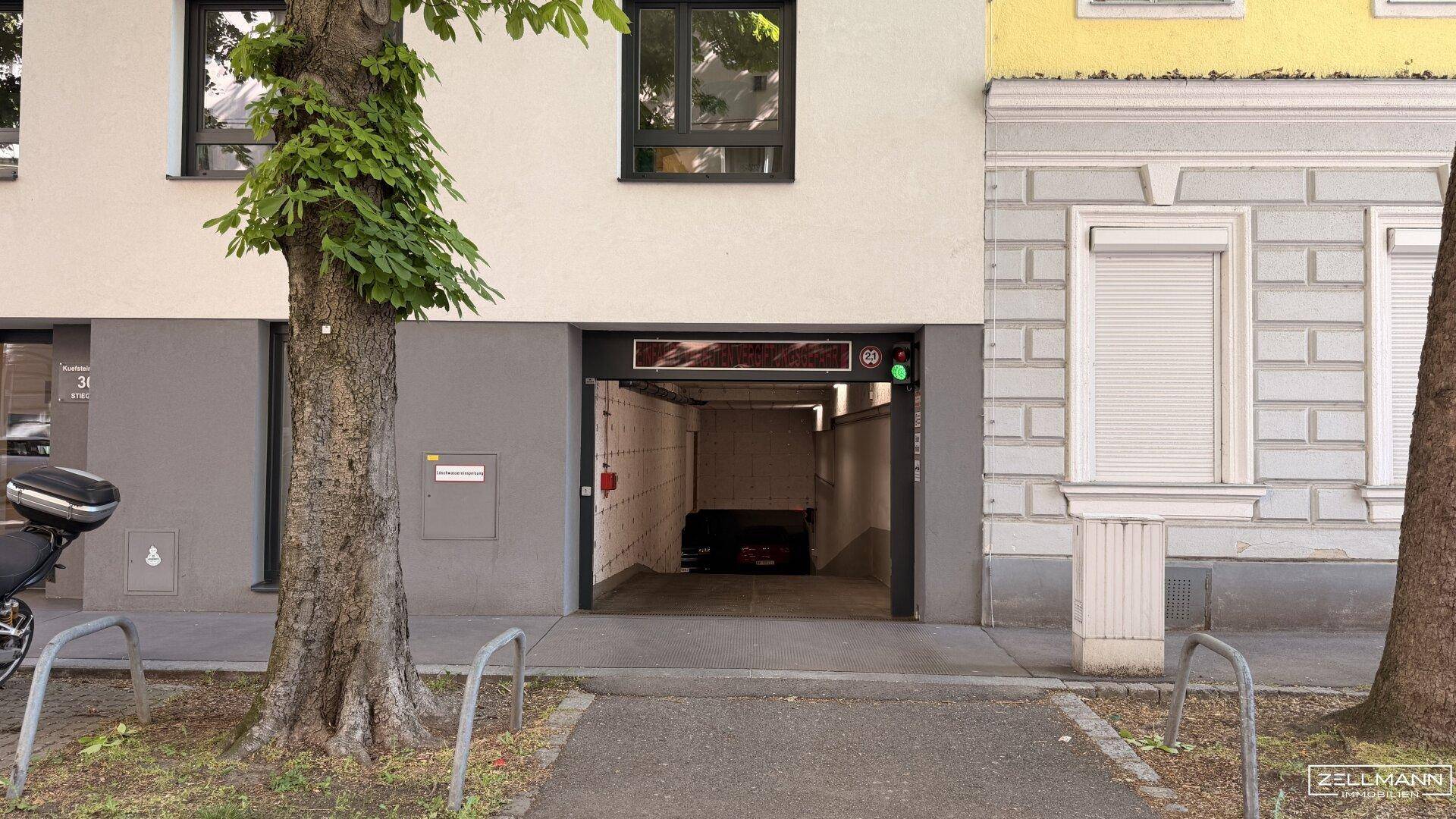 Fassade eines Gebäudes mit der Einfahrt zur Tiefgarage und einem Baum im Vordergrund.