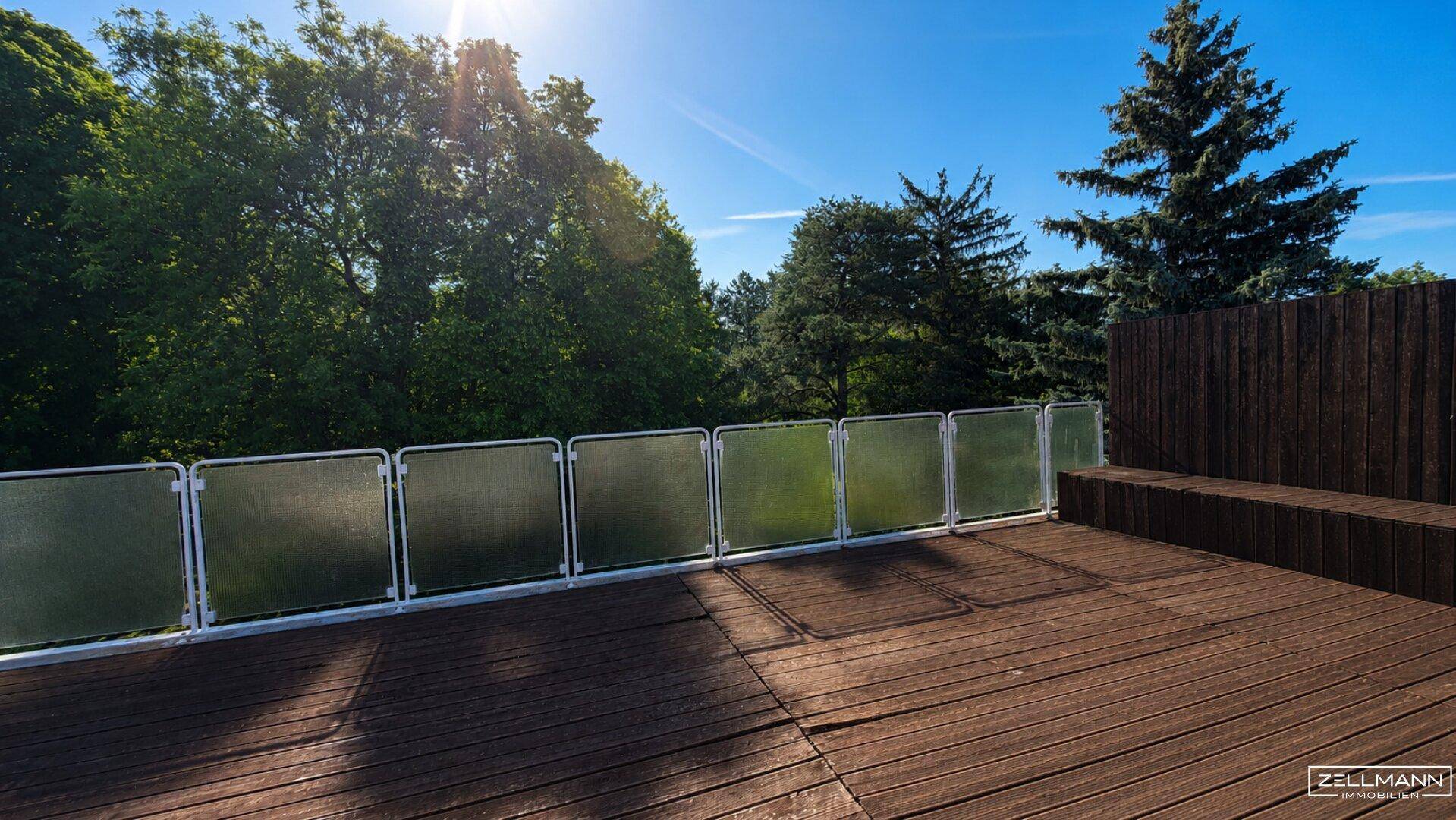 Große Dachterrasse mit Holzboden und Glasgeländer, umgeben von Bäumen unter blauem Himmel.