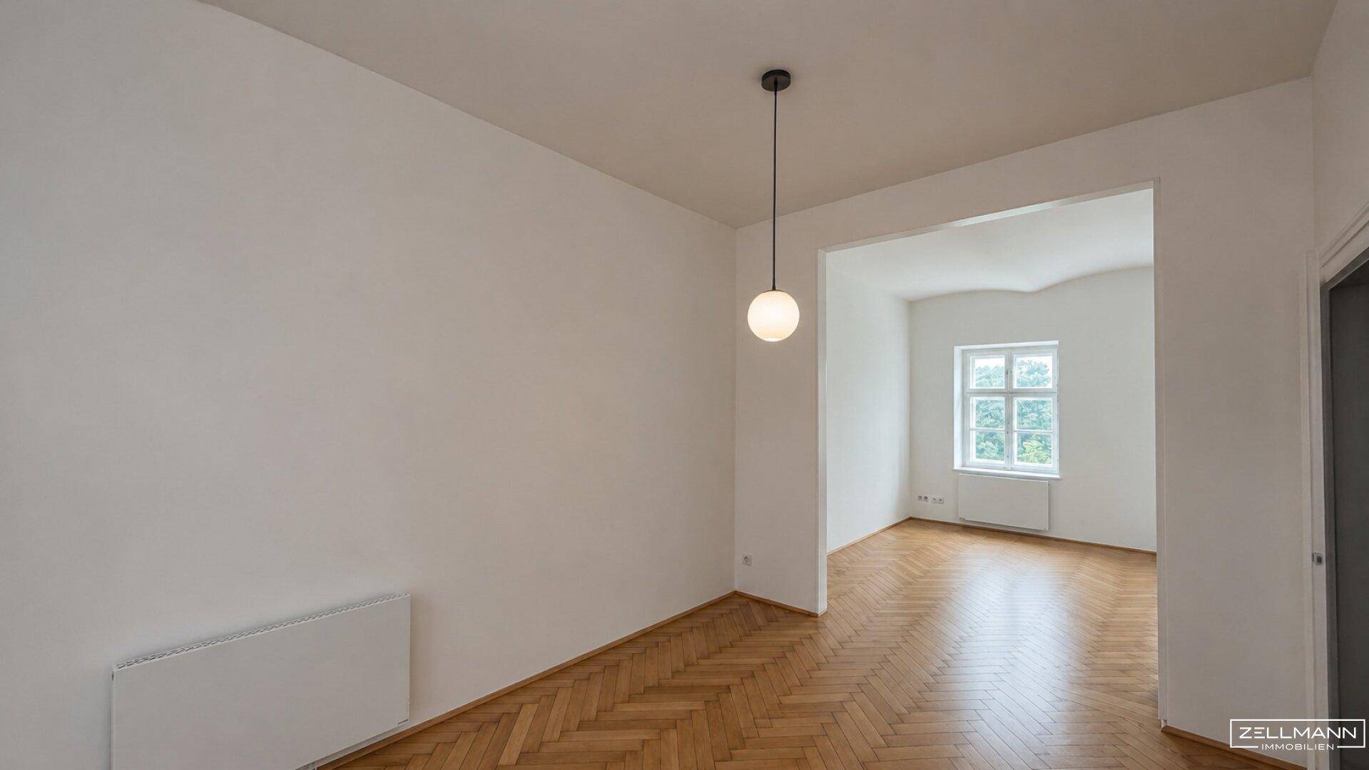 Geräumiger Wohnbereich mit Fischgrätparkett und Blick in einen weiteren Raum mit Fenster.