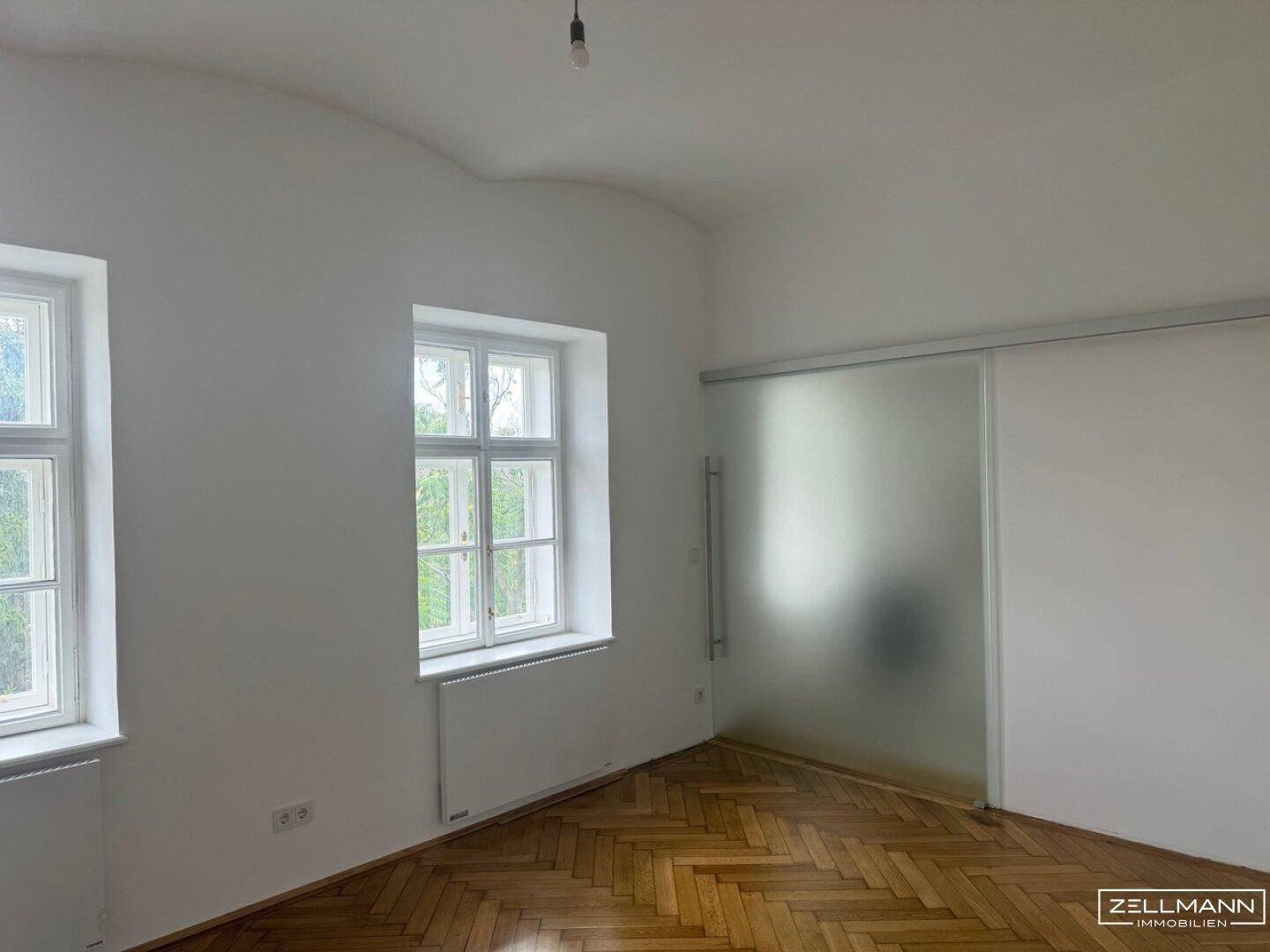 Heller Raum mit Fischgrätparkett und großen Fenstern, abgetrennt durch eine Glasschiebetür.