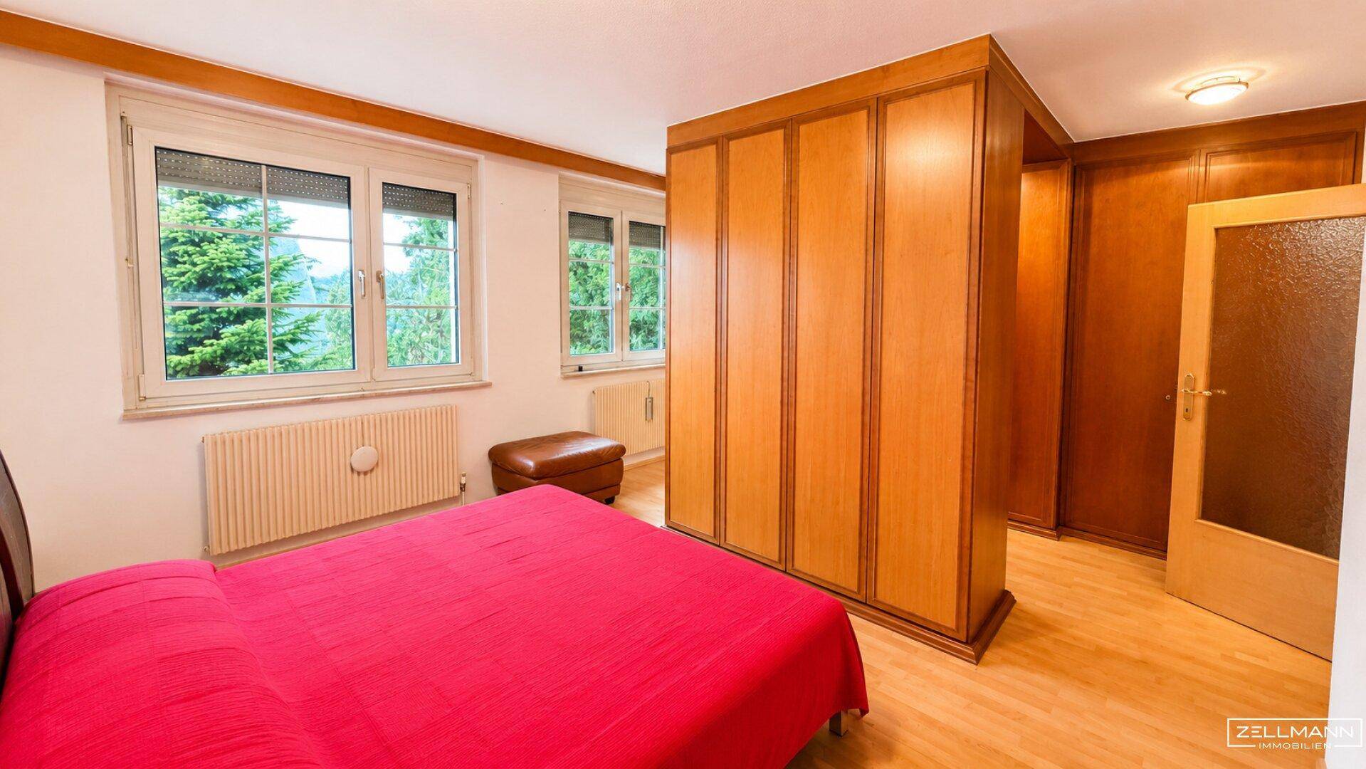 Schlafzimmer mit rotem Bett, Einbauschränken aus Holz und Fenstern mit Blick ins Grüne.