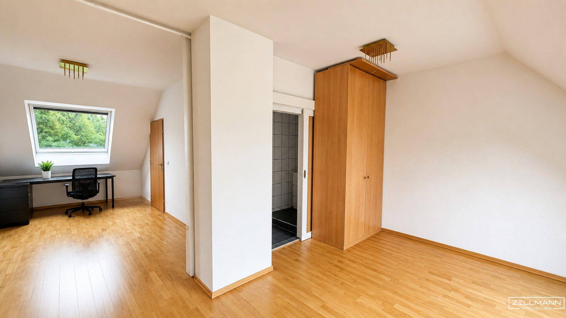 Helles Dachzimmer mit Schreibtisch, Einbauschrank und Zugang zu einem Badezimmer.