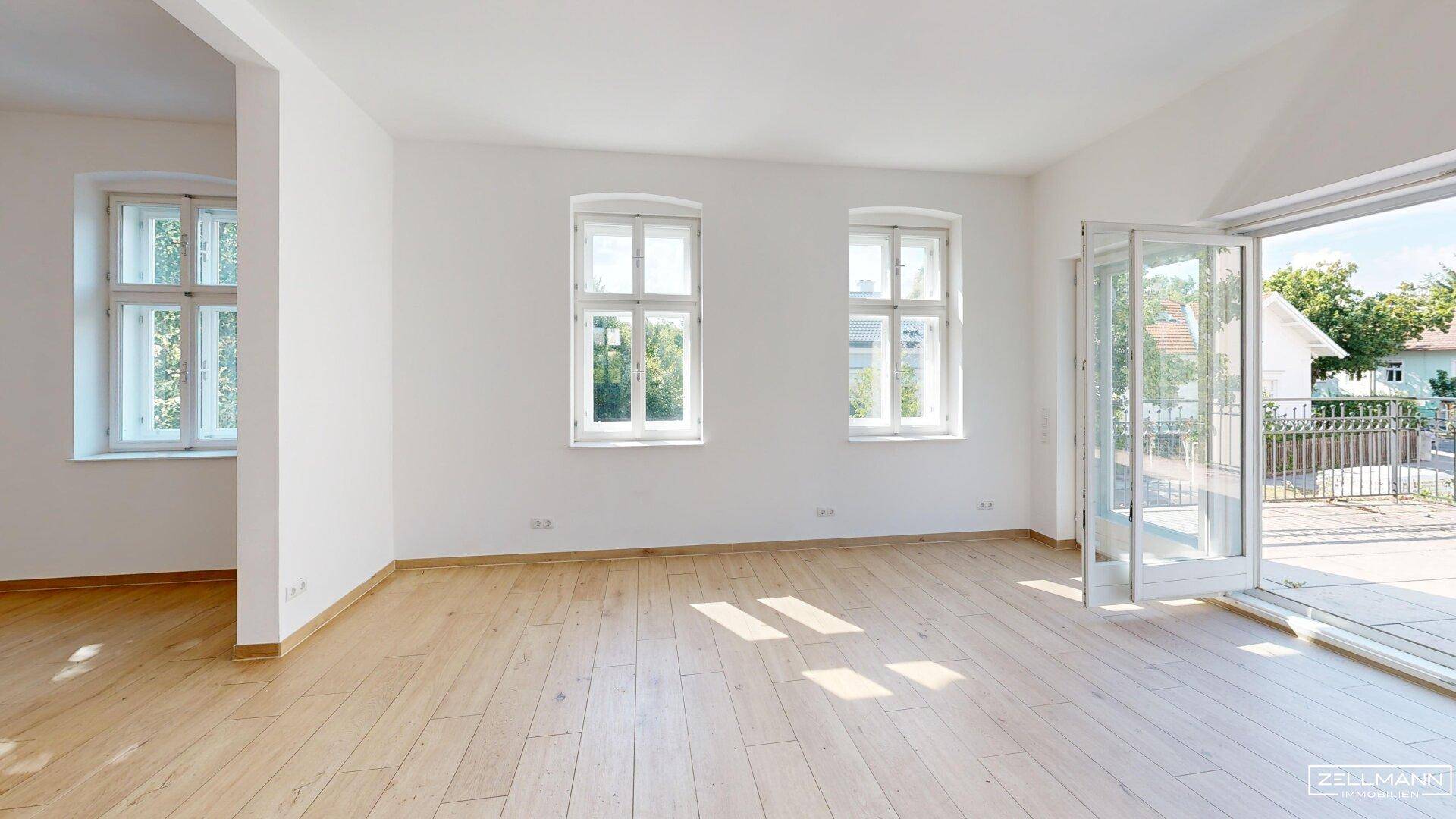 Geräumiges Zimmer mit Holzboden, großen Fenstern und Zugang zu einem Außenbereich.