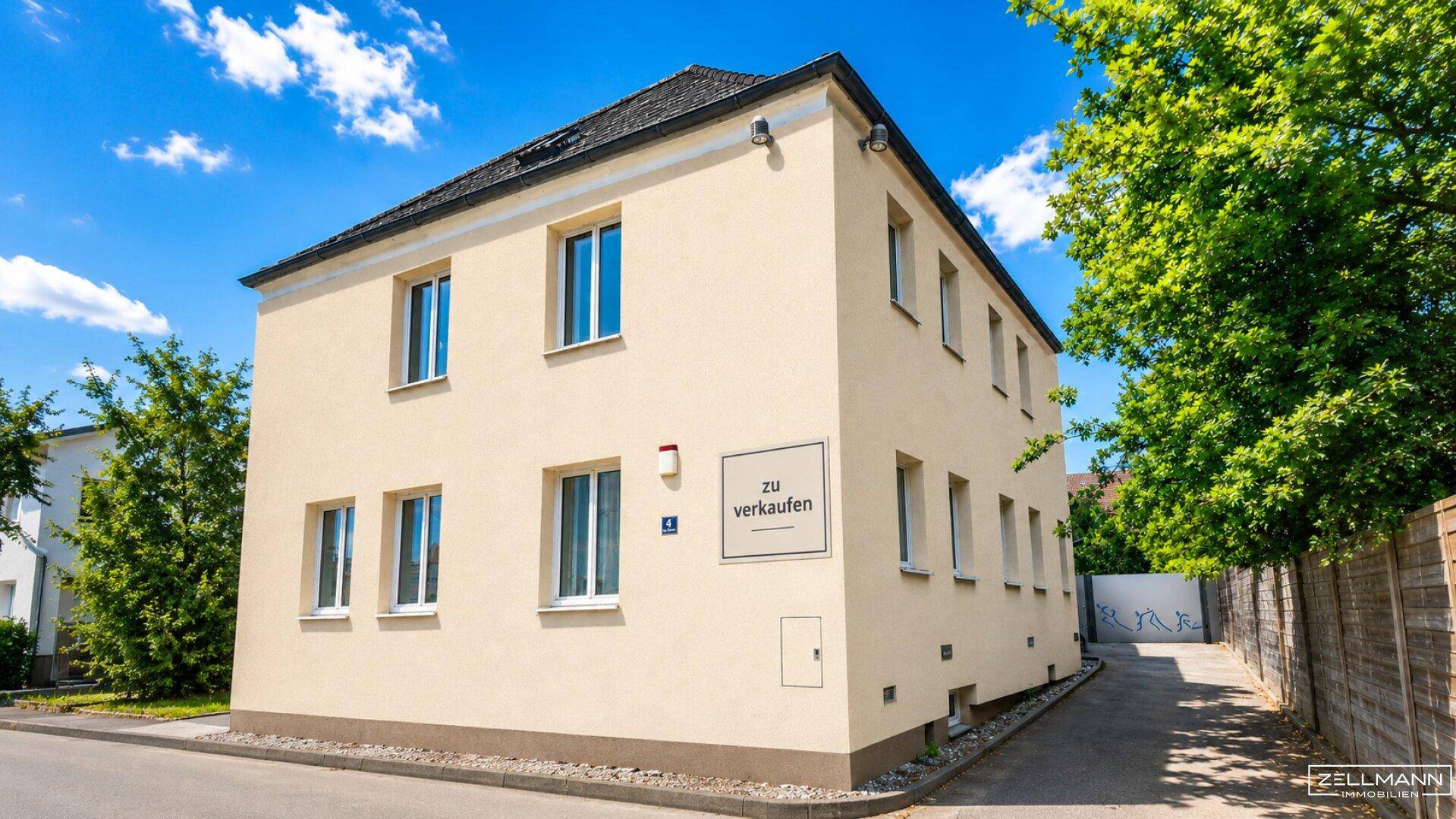 Freistehendes, hell gestrichenes Gebäude mit mehreren Fenstern und einem "zu verkaufen"-Schild an der Fassade.