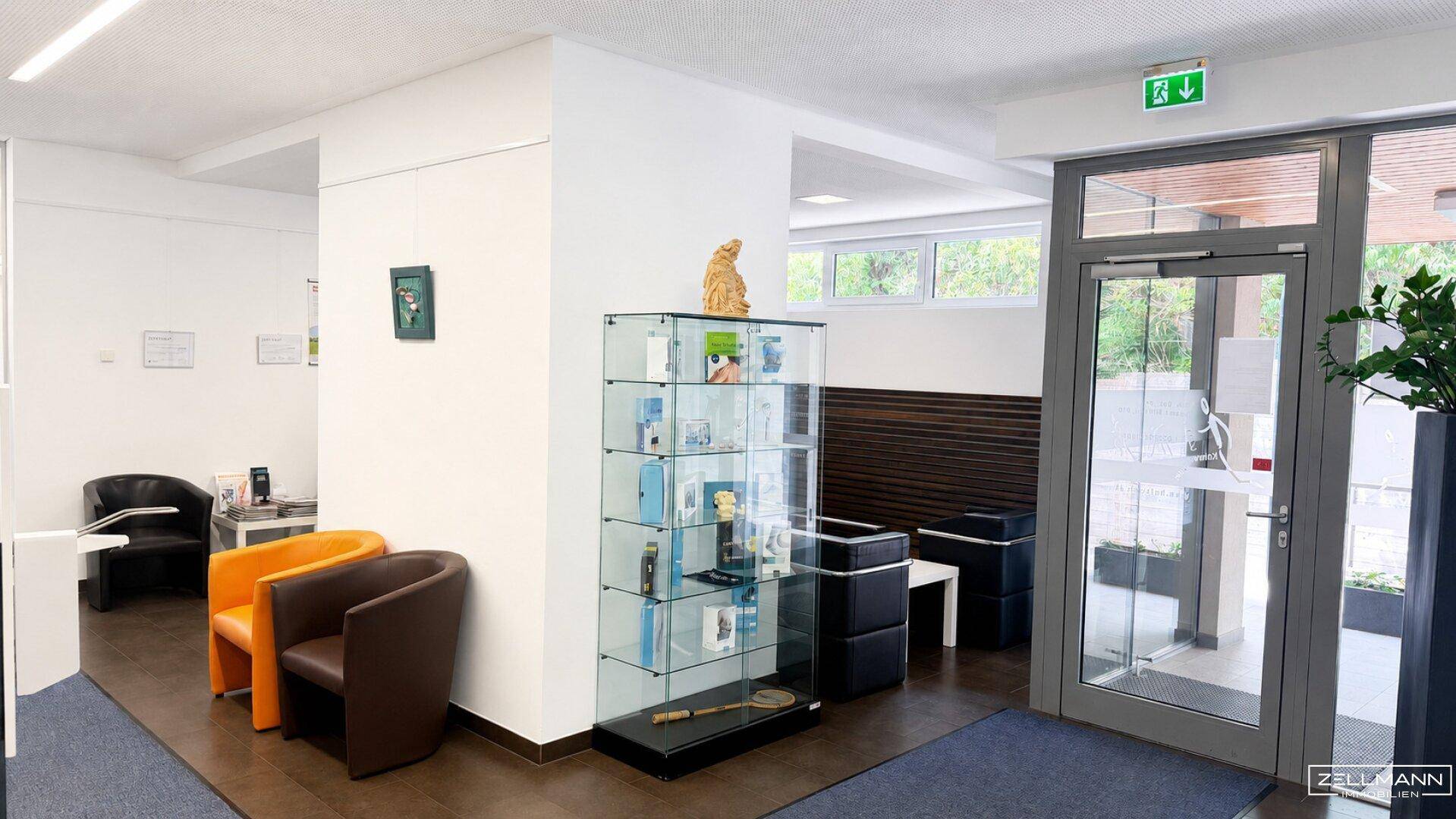 Heller Empfangsbereich mit Glasvitrine, modernen Sitzgelegenheiten und großem Eingang.