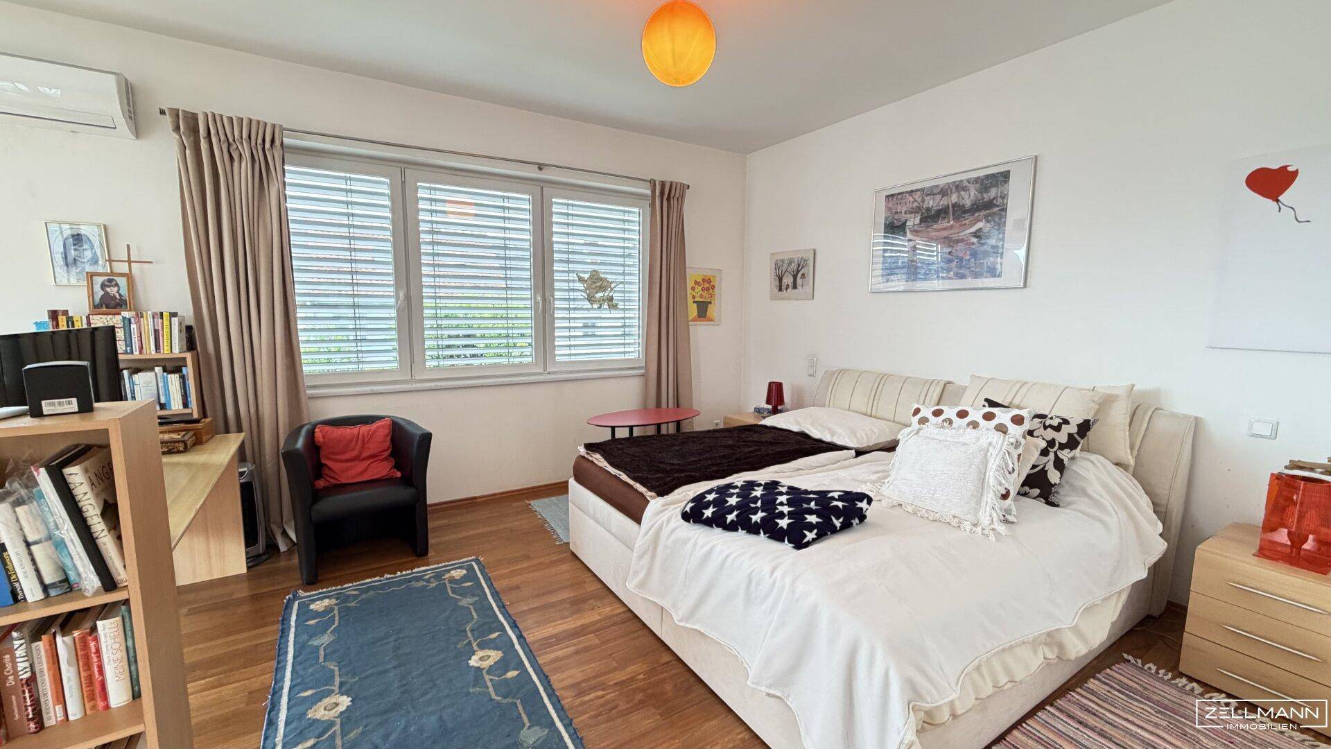 Gemütliches Schlafzimmer mit großem Bett, Bücherregal und Fenster mit Jalousien.