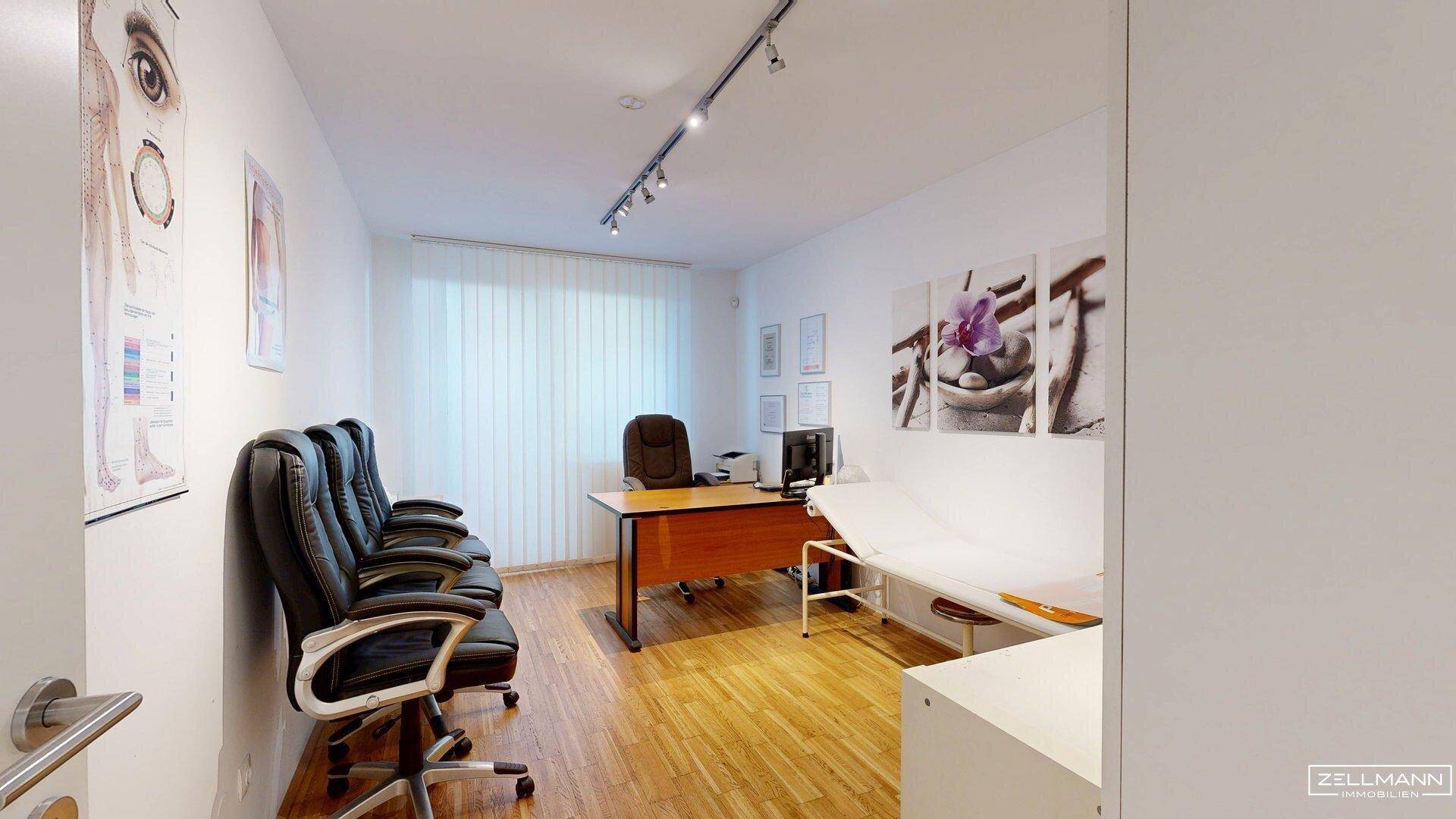 Helles Behandlungszimmer mit Schreibtisch, Untersuchungsliege und zusätzlichen Sitzgelegenheiten für Besucher.