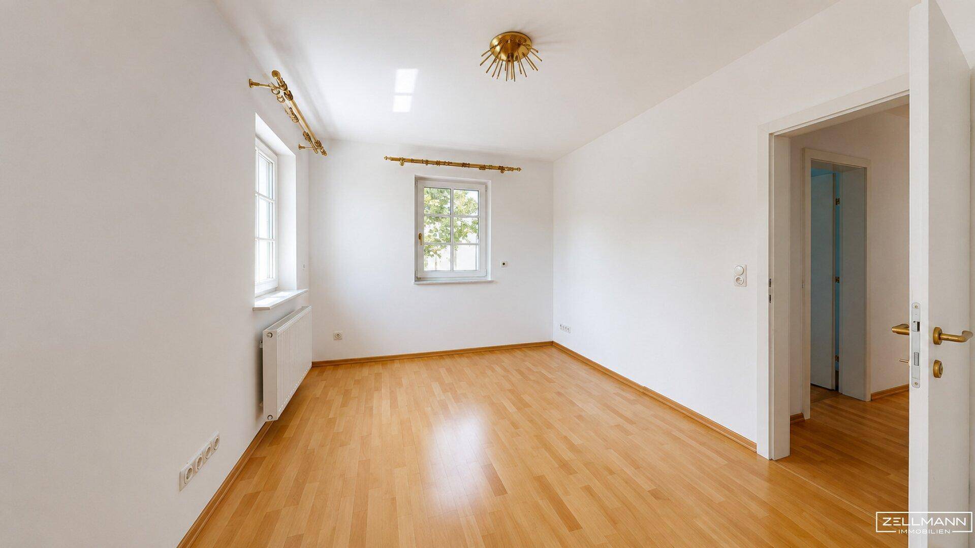 Leeres Zimmer mit hellem Holzfußboden, weißen Wänden und zwei Fenstern mit Blick ins Grüne.
