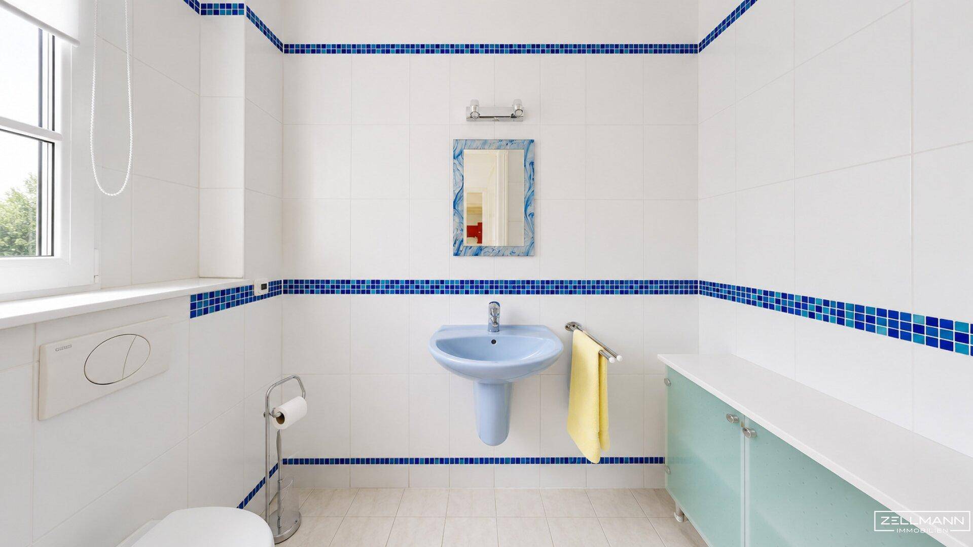 Badezimmer mit weißen Fliesen, blauen Akzenten, hellblauem Waschbecken und Fenster.