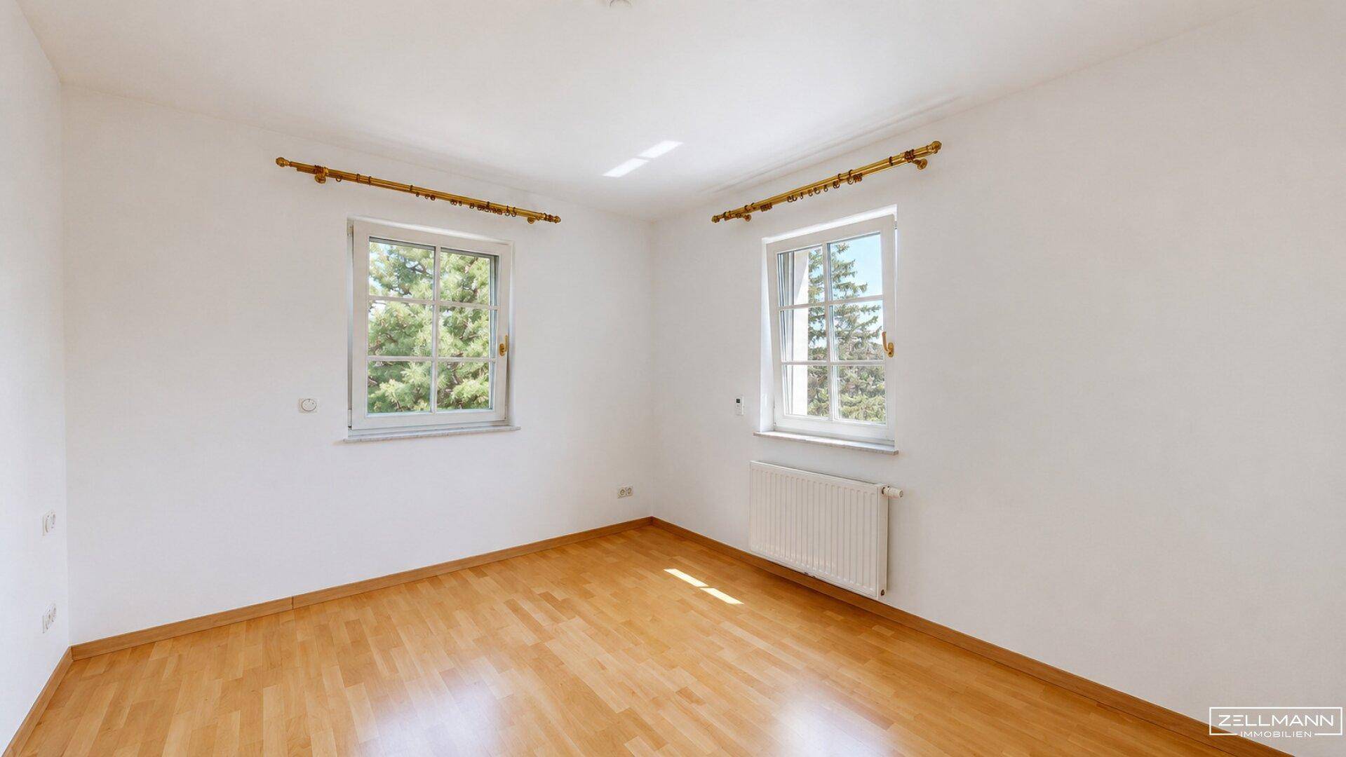 Geräumiges Zimmer mit hellem Holzfußboden, weißen Wänden und zwei großen Fenstern.