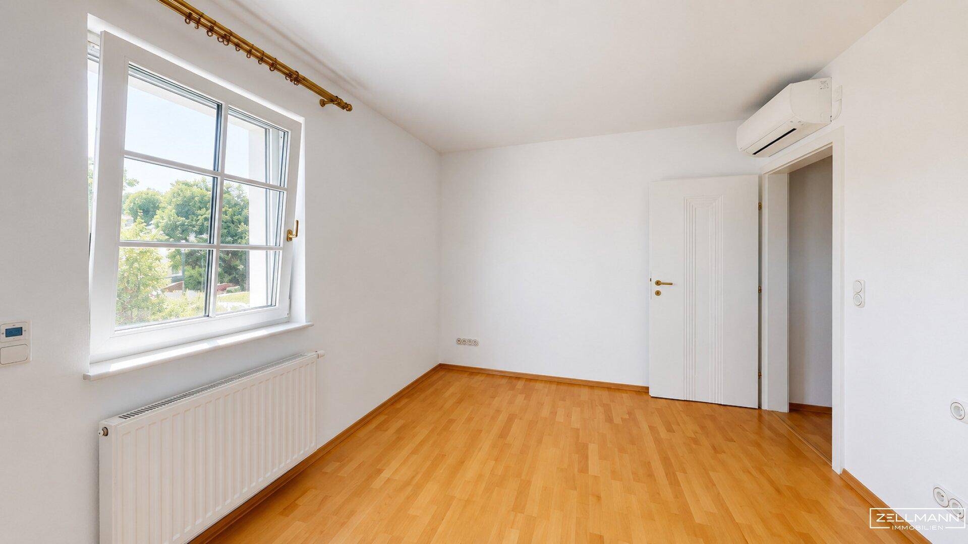 Helles Zimmer mit Holzfußboden, Klimaanlage und Fenster mit Ausblick ins Grüne.