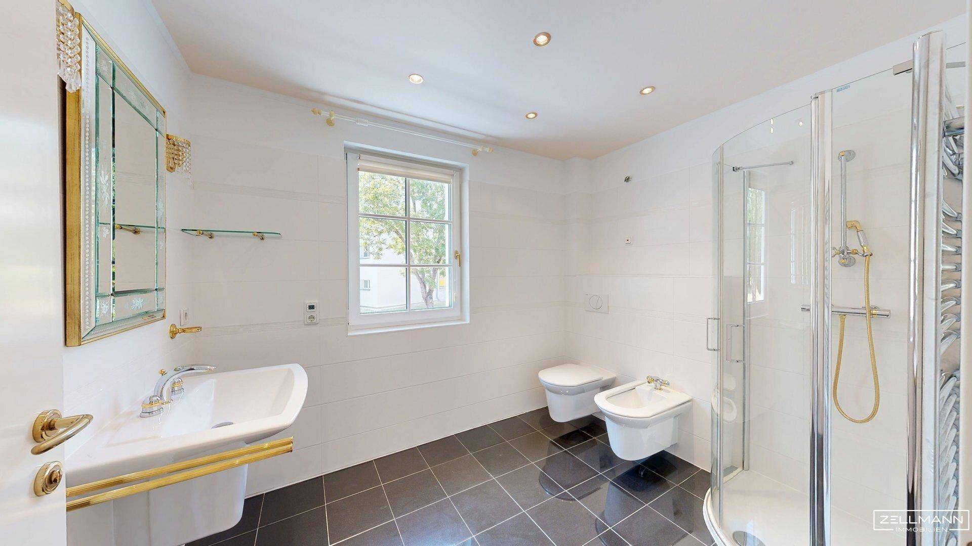 Helles Badezimmer mit Waschtisch, Toilette, Bidet, Glasdusche und Fenster.
