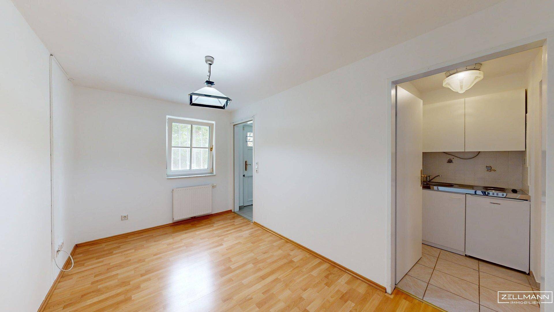 Wohnraum mit hellem Holzfußboden, Fenster und integrierter kleiner Küchenzeile.