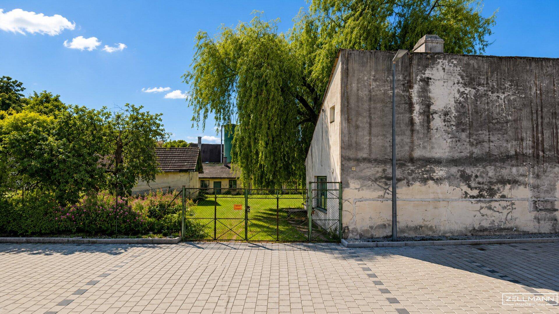 Blick auf ein eingezäuntes Grundstück mit einem alten Gebäude und üppiger Vegetation unter blauem Himmel.