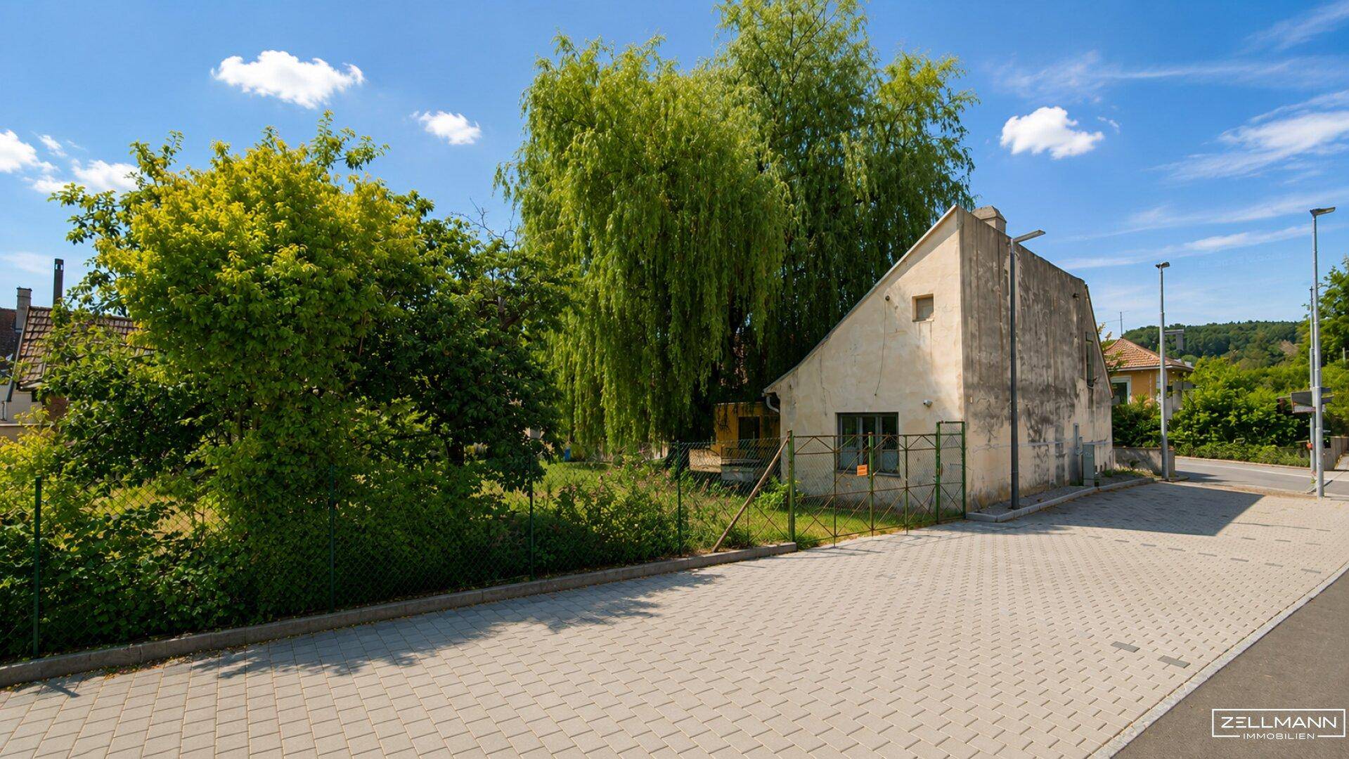 Seitenansicht des alten Gebäudes mit großem Baum, gepflastertem Vorplatz und grüner Umgebung.
