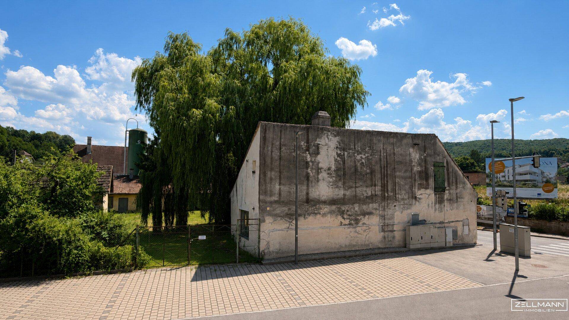 Außenansicht eines älteren Gebäudes mit verputzter Fassade, umgeben von Bäumen und einem gepflasterten Bereich.