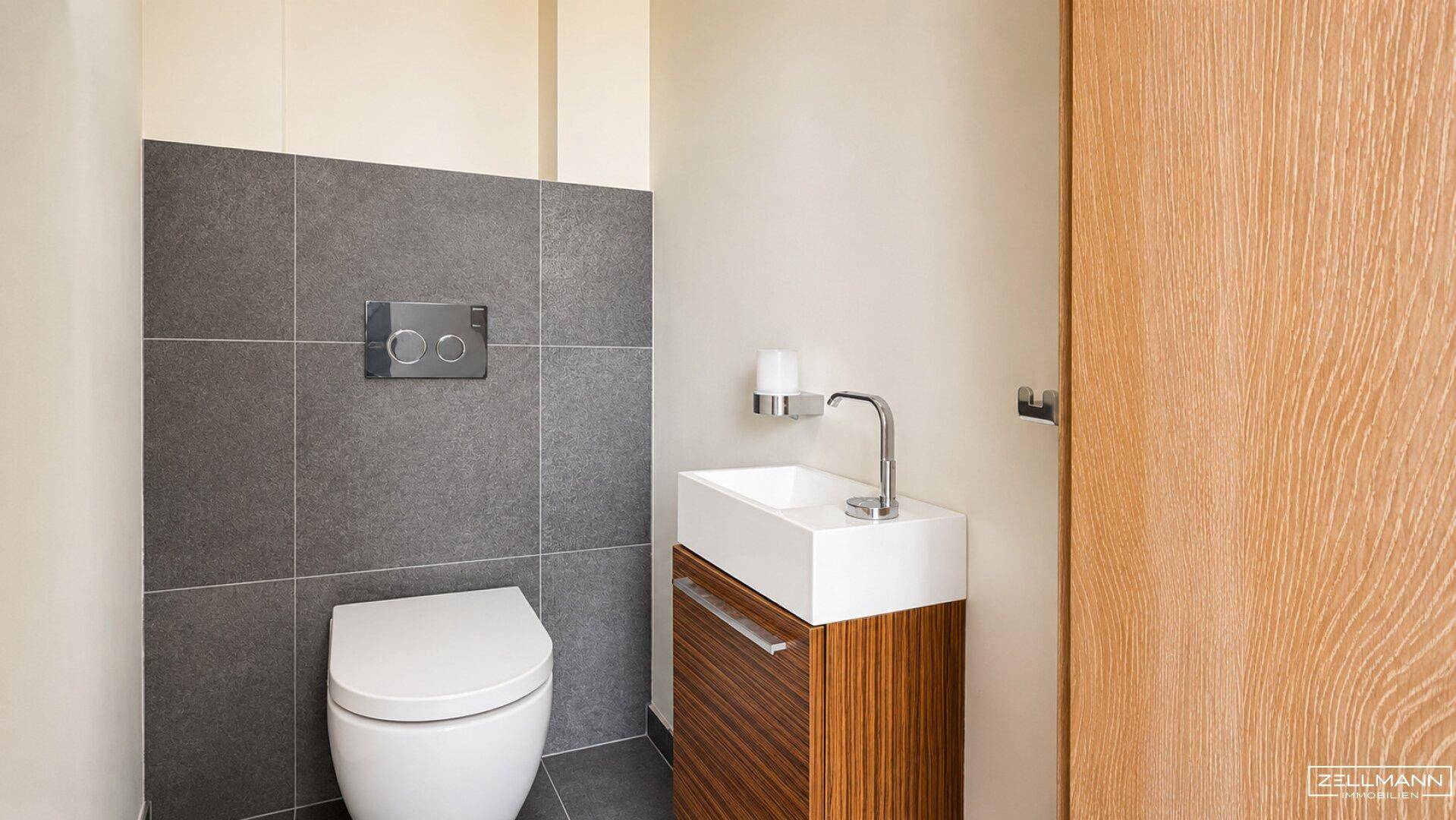 Modernes Gäste-WC mit Wand-WC, kleinem Waschbecken und grauen Wandfliesen.