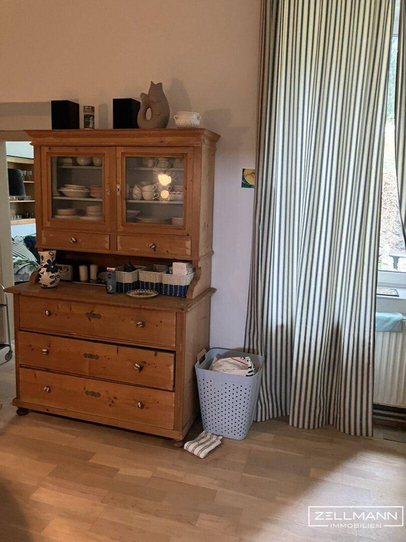 Innenraum mit hellem Holzboden, einem antiken Holzschrank und gestreiften Vorhängen am Fenster.
