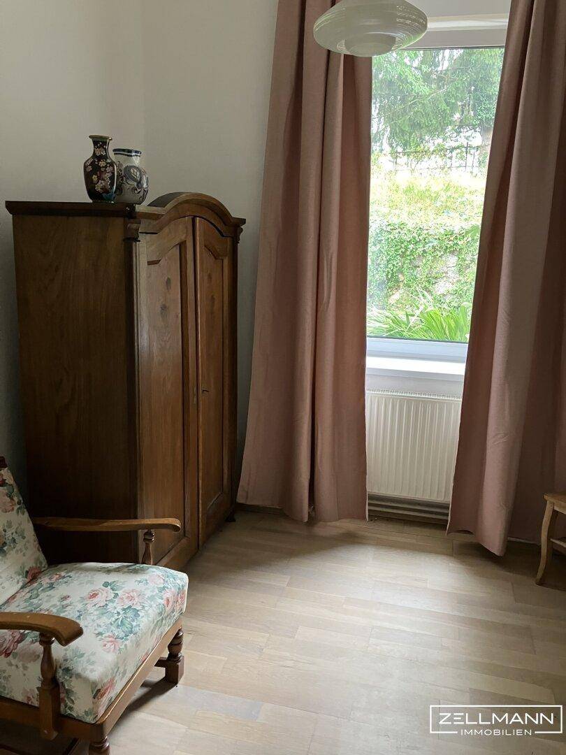 Schlafzimmer mit hellem Holzboden, einem antiken Holzschrank und Vorhängen am Fenster.