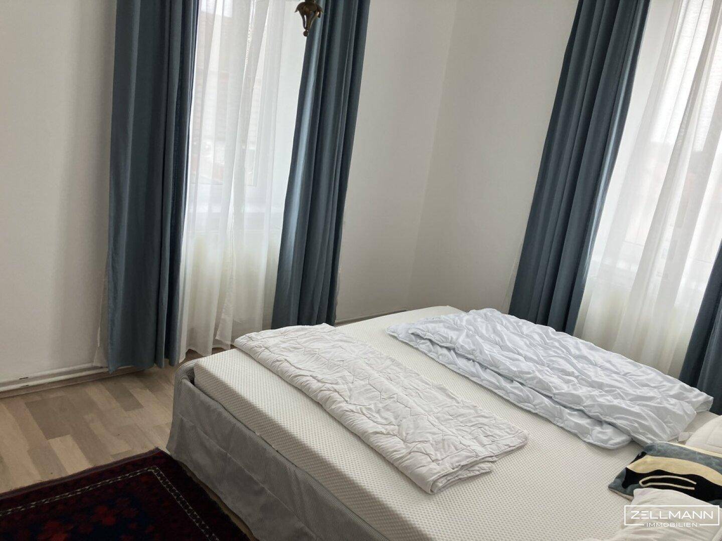 Schlafzimmer mit Doppelbett, hellem Holzboden und blauen Vorhängen an den Fenstern.
