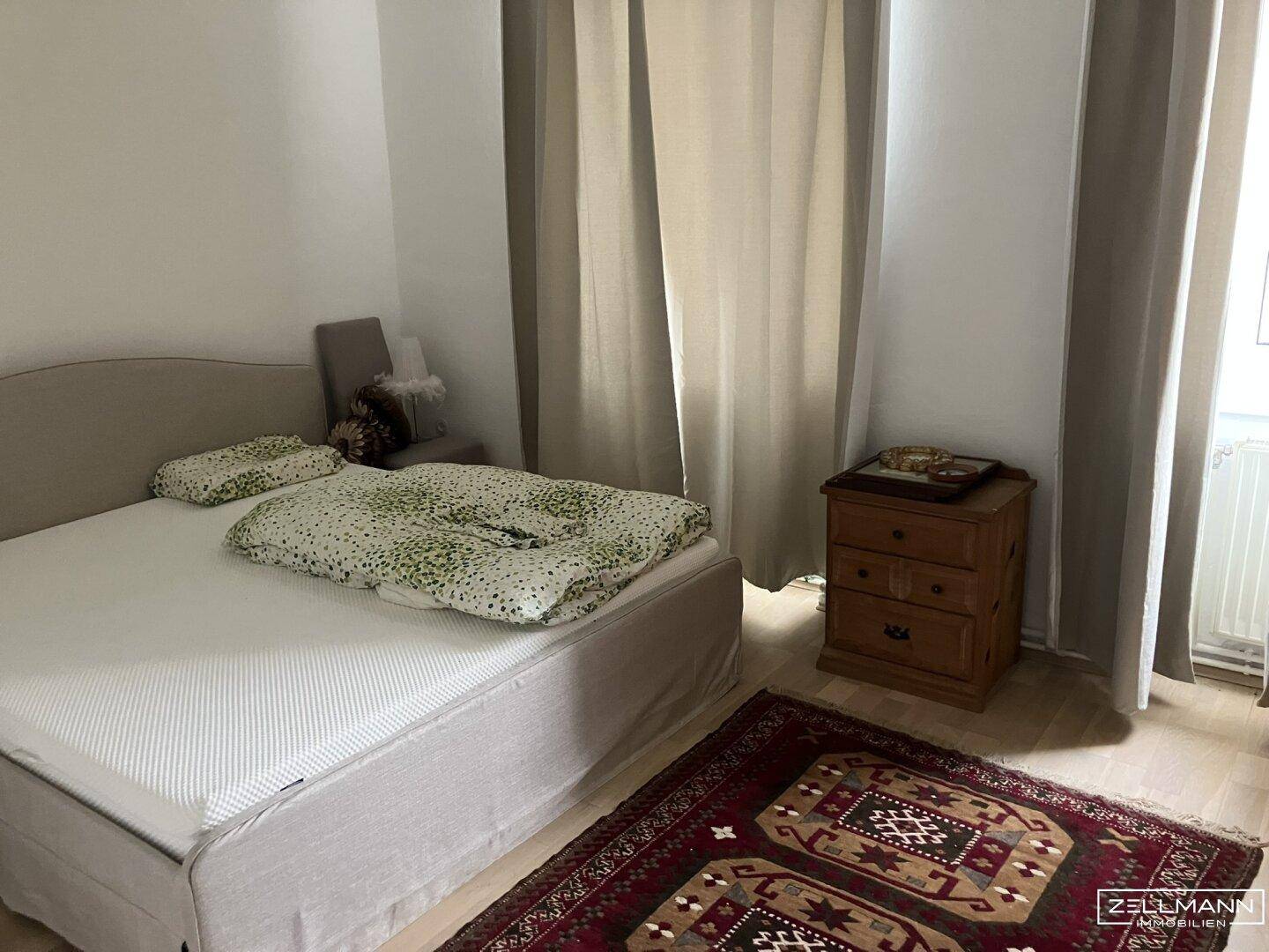 Schlafzimmer mit Bett, Nachttisch und einem Teppich auf dem hellen Holzboden.
