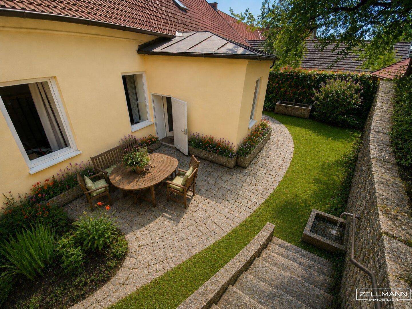 Sonniger Außenbereich mit gepflasterter Terrasse, Gartenmöbeln und gepflegtem Rasen.