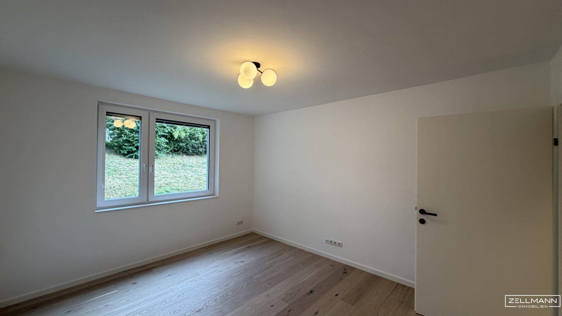 Leeres Zimmer mit hellem Holzboden und Fenster, das viel Tageslicht hereinlässt.
