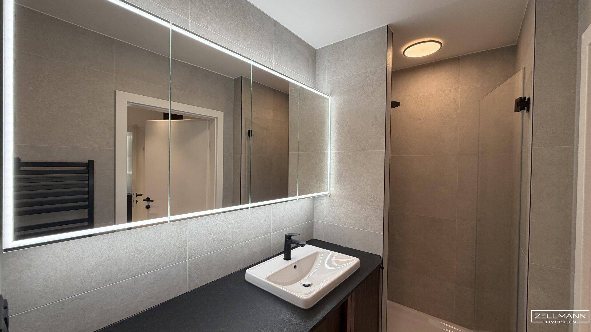 Modernes Badezimmer mit großformatigen grauen Fliesen, beleuchtetem Spiegelschrank und Einzelwaschbecken.
