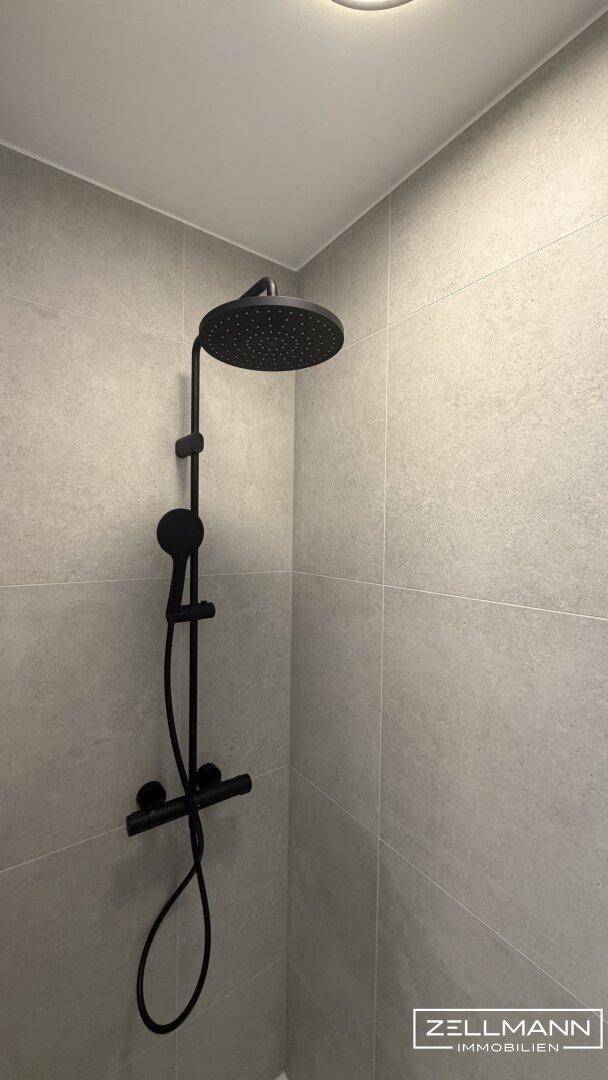 Moderne Duschkabine mit schwarzer Regendusche und grauen Wandfliesen.