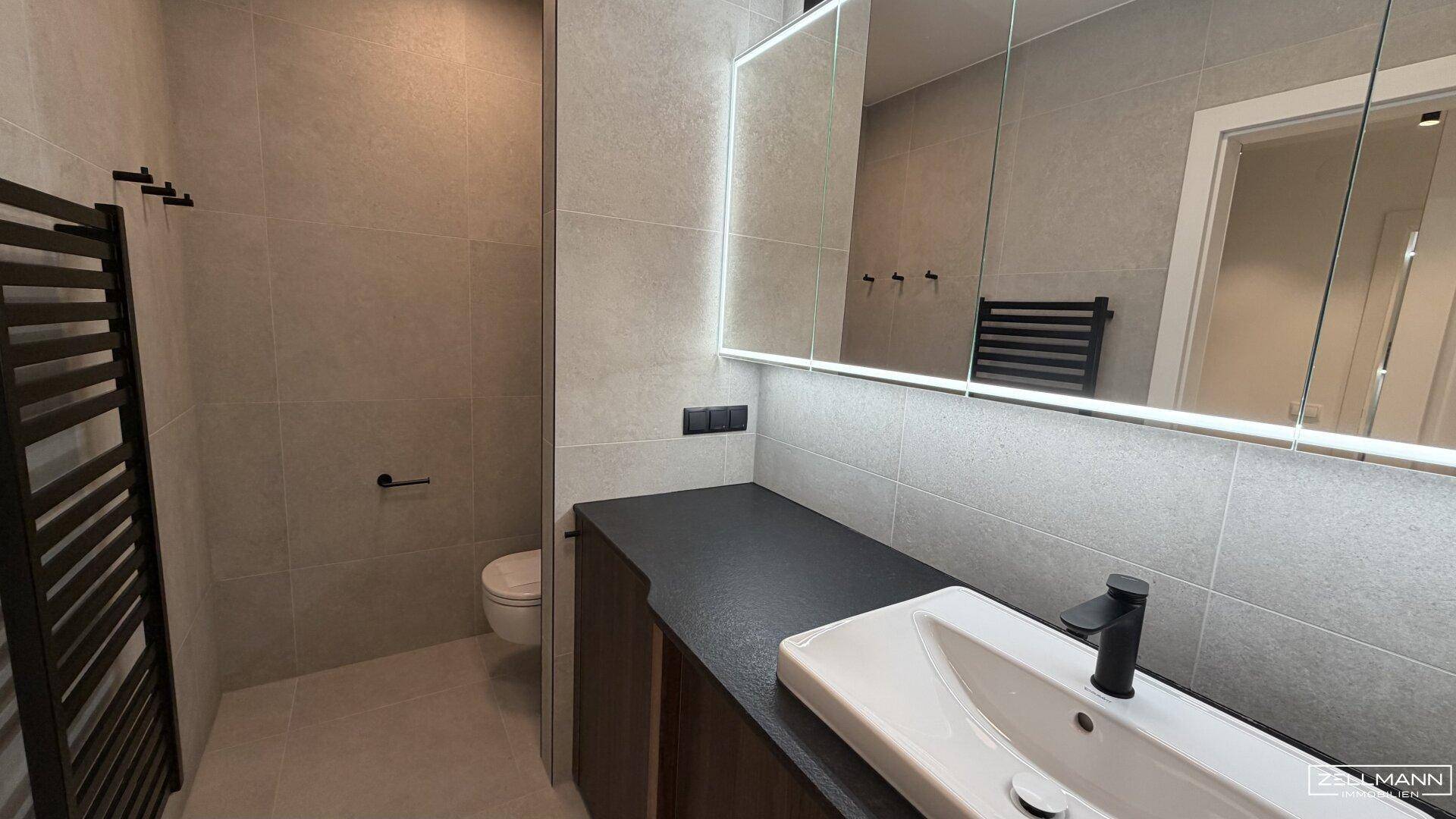 Modernes Badezimmer mit grauen Fliesen, wandhängendem WC und beheiztem Handtuchhalter.