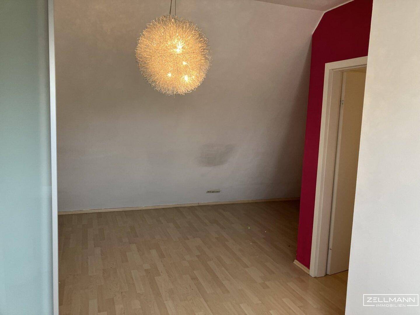 Geräumiges Dachzimmer mit schrägen Wänden, Laminatboden und auffälliger Deckenleuchte.