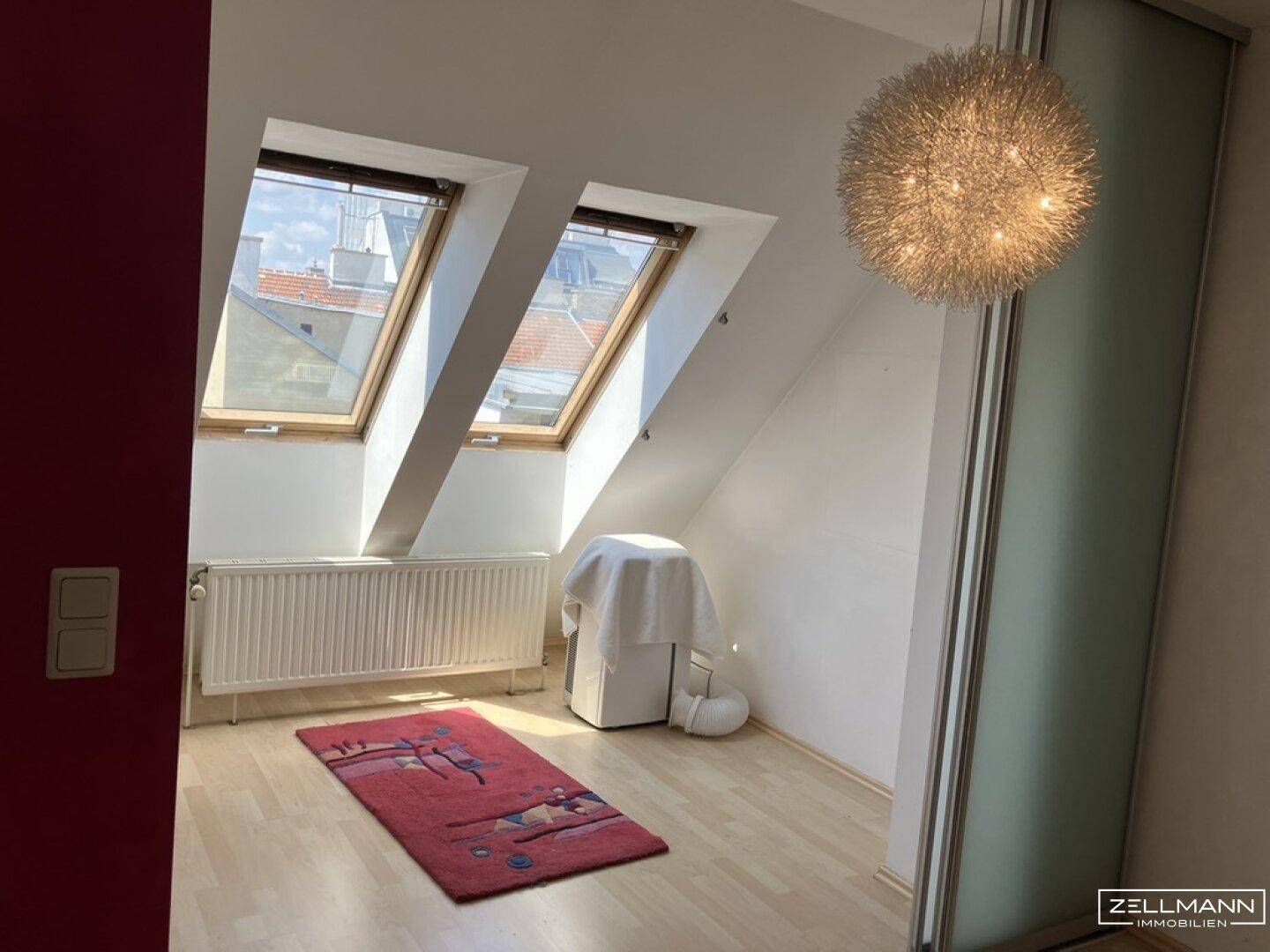 Helles Dachzimmer mit zwei Dachfenstern, Holzfußboden und moderner Deckenleuchte.