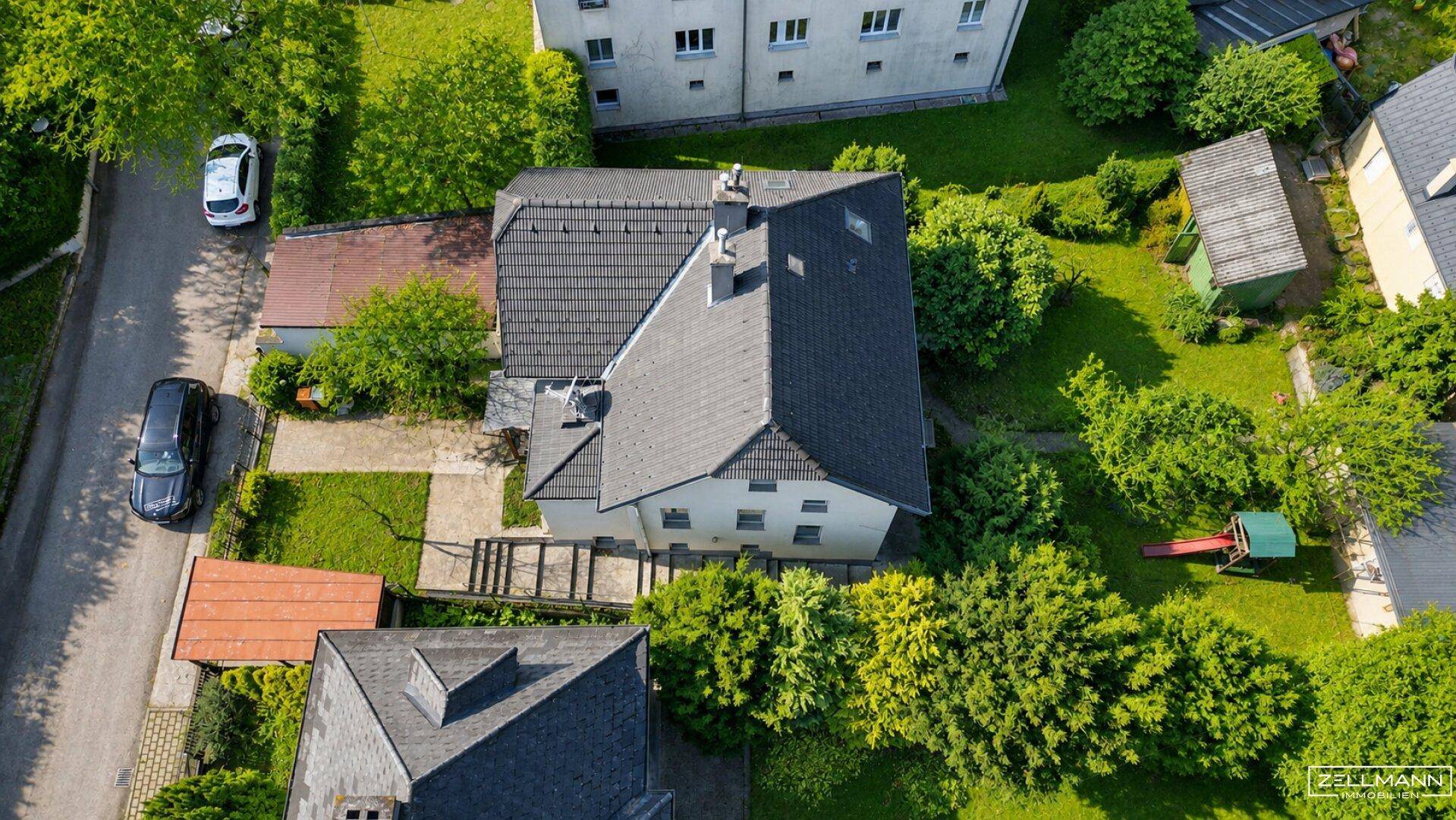 Luftaufnahme des Hauses mit dunklem Ziegeldach, großem Garten und angrenzender Straße.