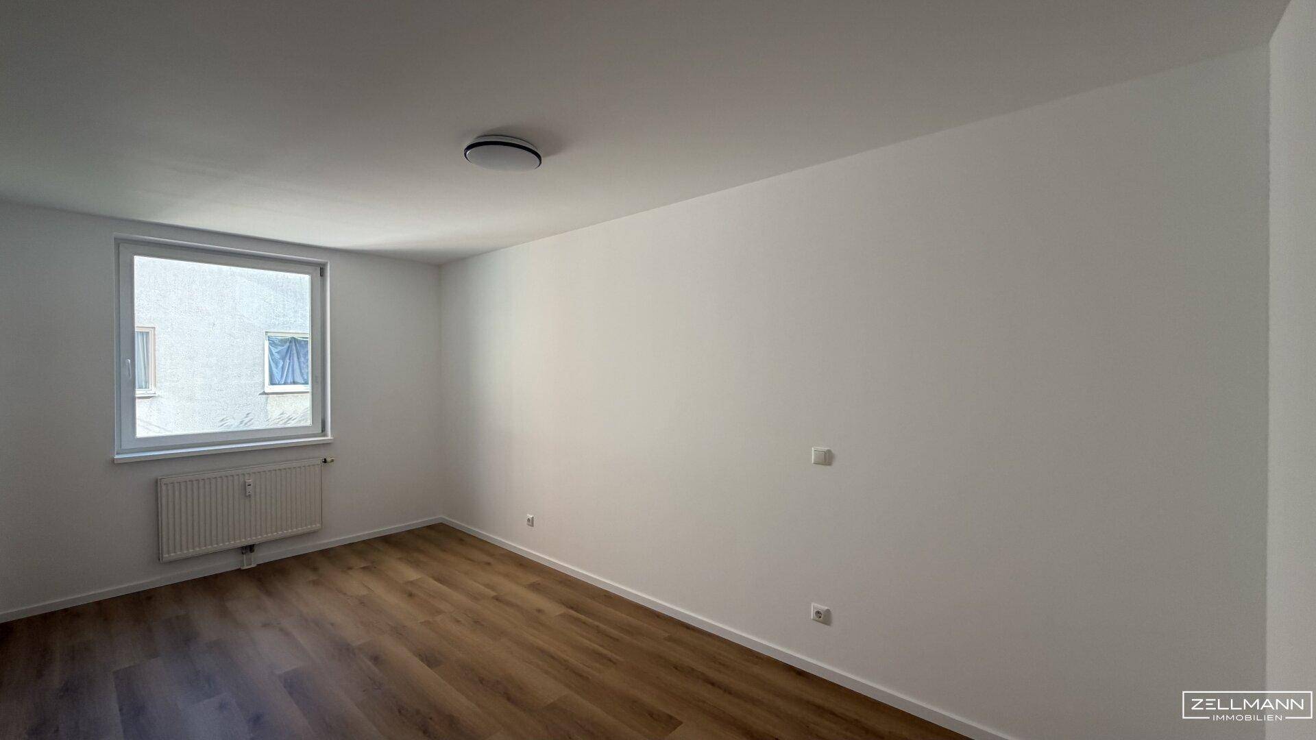 Helles Zimmer mit Holzboden, weißen Wänden und einem Fenster mit Blick nach draußen.