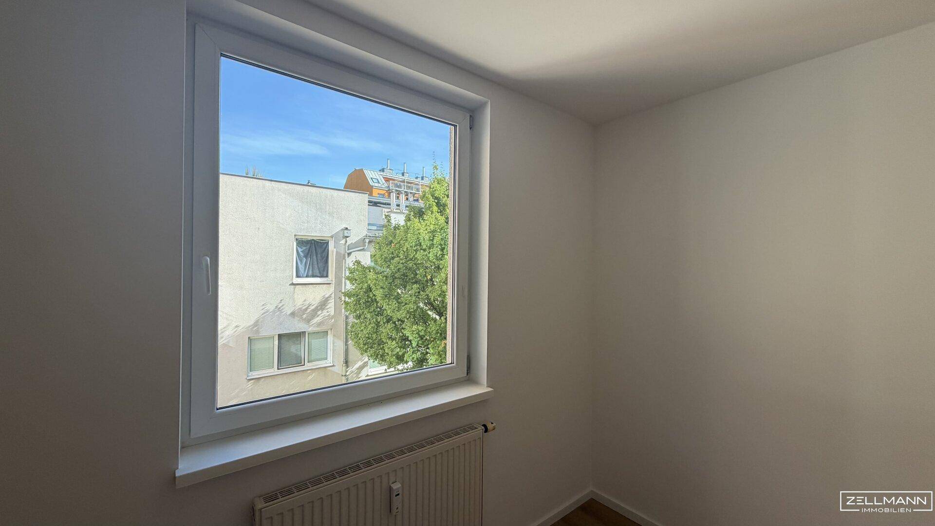 Freundliches Zimmer mit Holzboden und einem Fenster, das einen Blick auf die umliegenden Gebäude bietet.