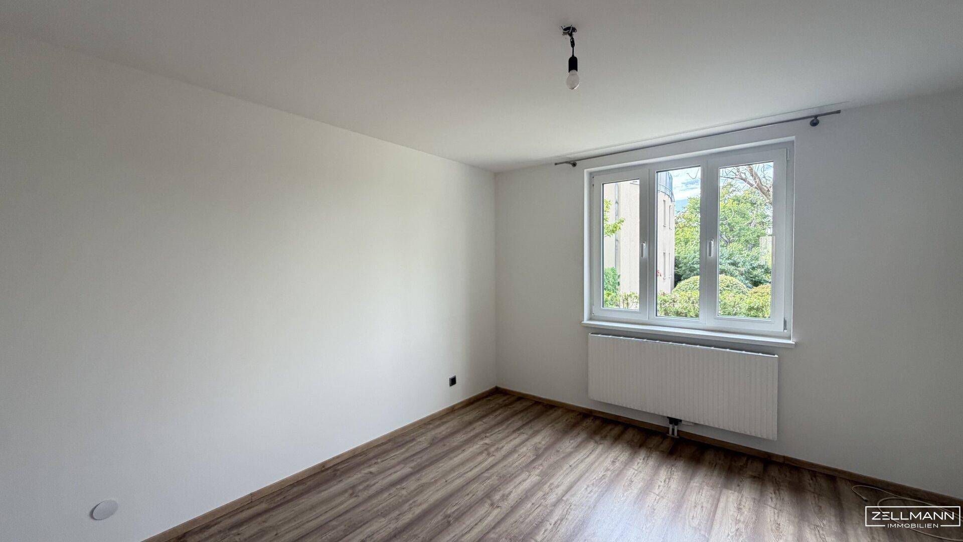 Schlafzimmer mit Holzboden und großem Fenster, das viel Tageslicht hereinlässt.