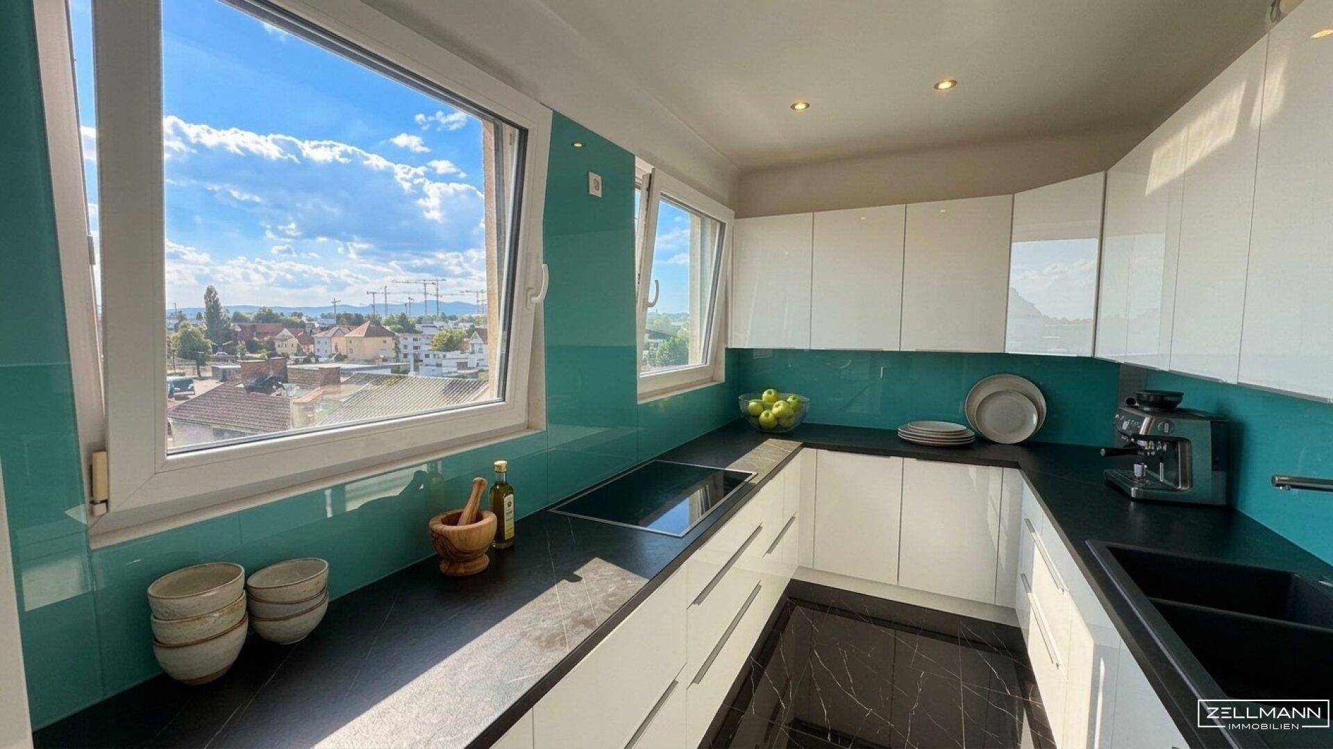 Moderne Küche mit weißen Hochglanzfronten, türkiser Rückwand und weitem Blick aus dem Fenster.