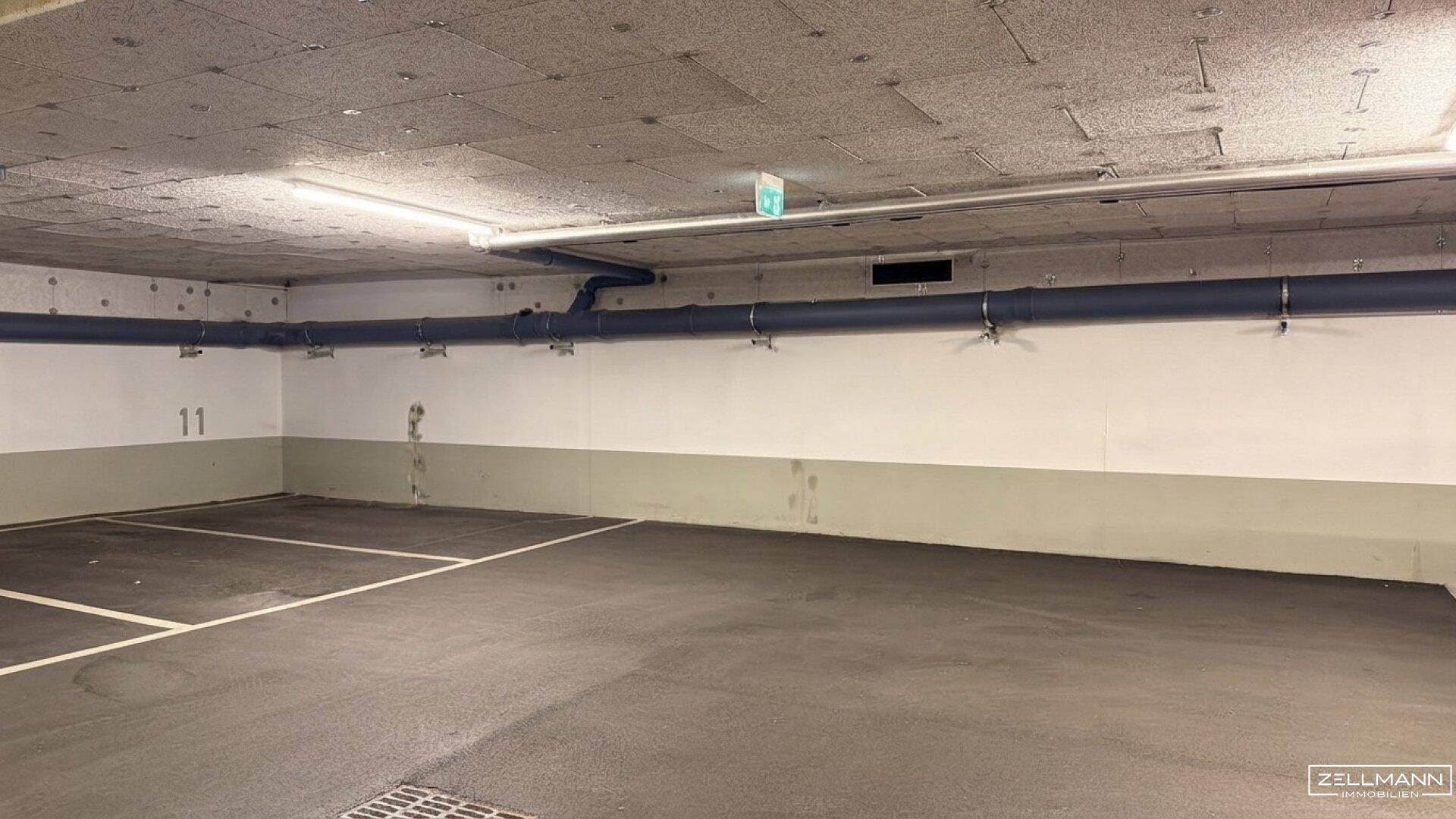 Weitere Ansicht der Tiefgarage mit markierten Stellplätzen, Betonwänden und Deckenbeleuchtung.