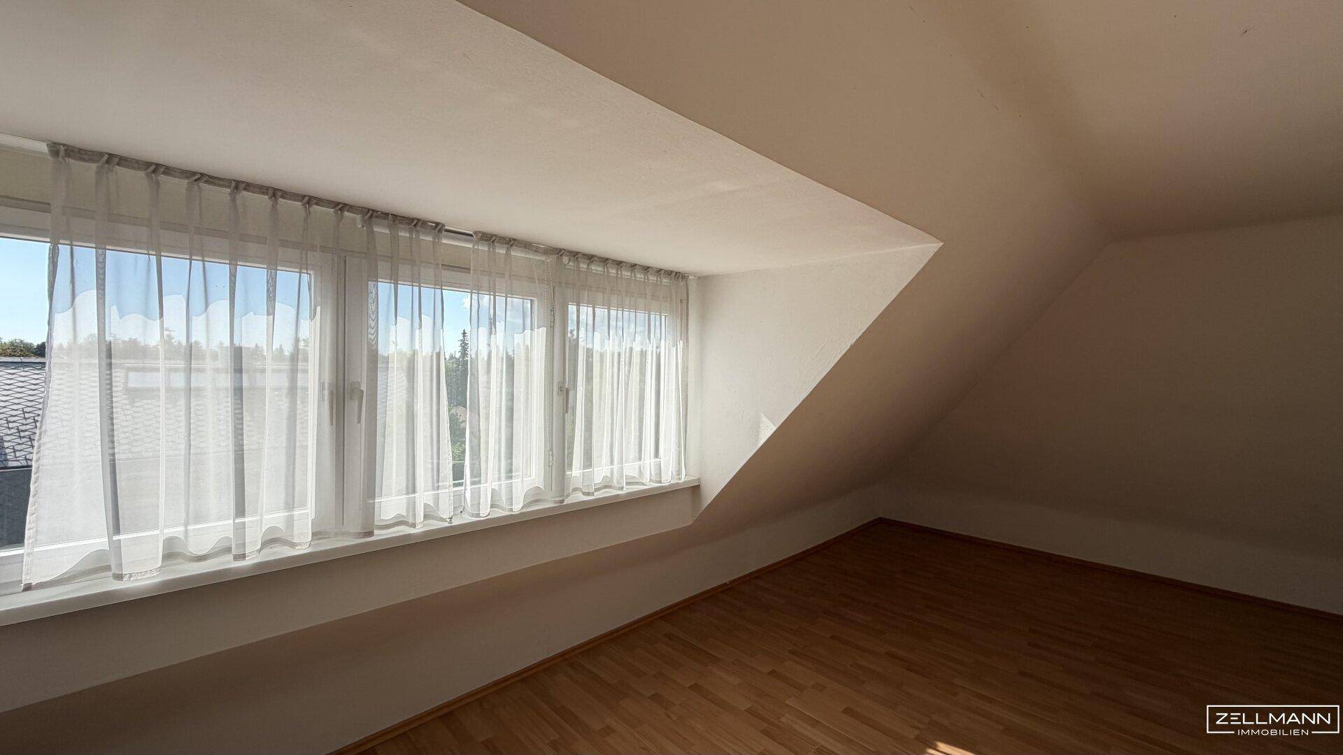 Gemütliches Dachzimmer mit Schrägdecke, Holzfußboden und breitem Fenster für viel Tageslicht.