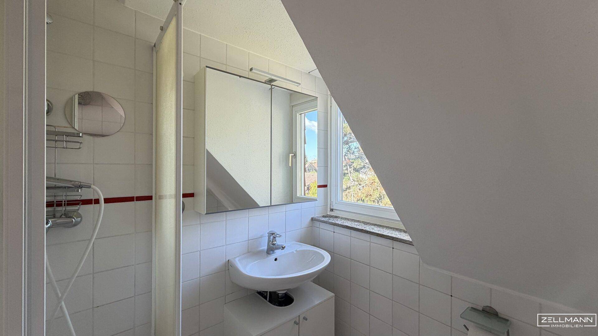 Helles Badezimmer mit Dusche, Waschbecken, Spiegelschrank und Fenster in der Dachschräge.