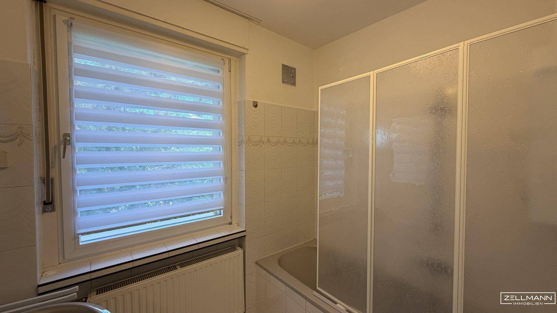 Badezimmer mit Badewanne und Duschabtrennung, Fenster mit Jalousie und hellen Fliesen.