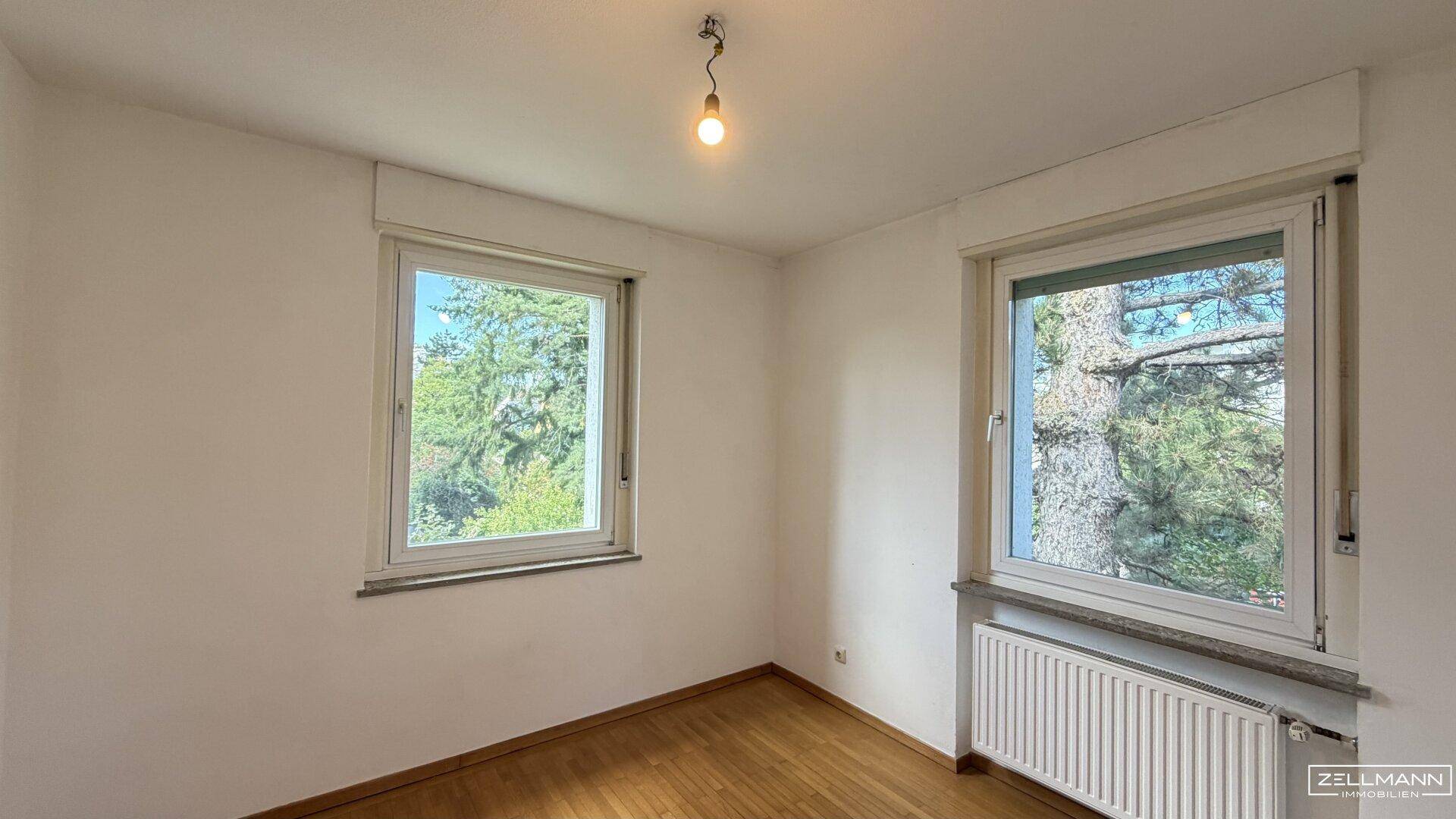 Helles Schlafzimmer mit Parkettboden und zwei Fenstern, die einen Blick ins Grüne bieten.