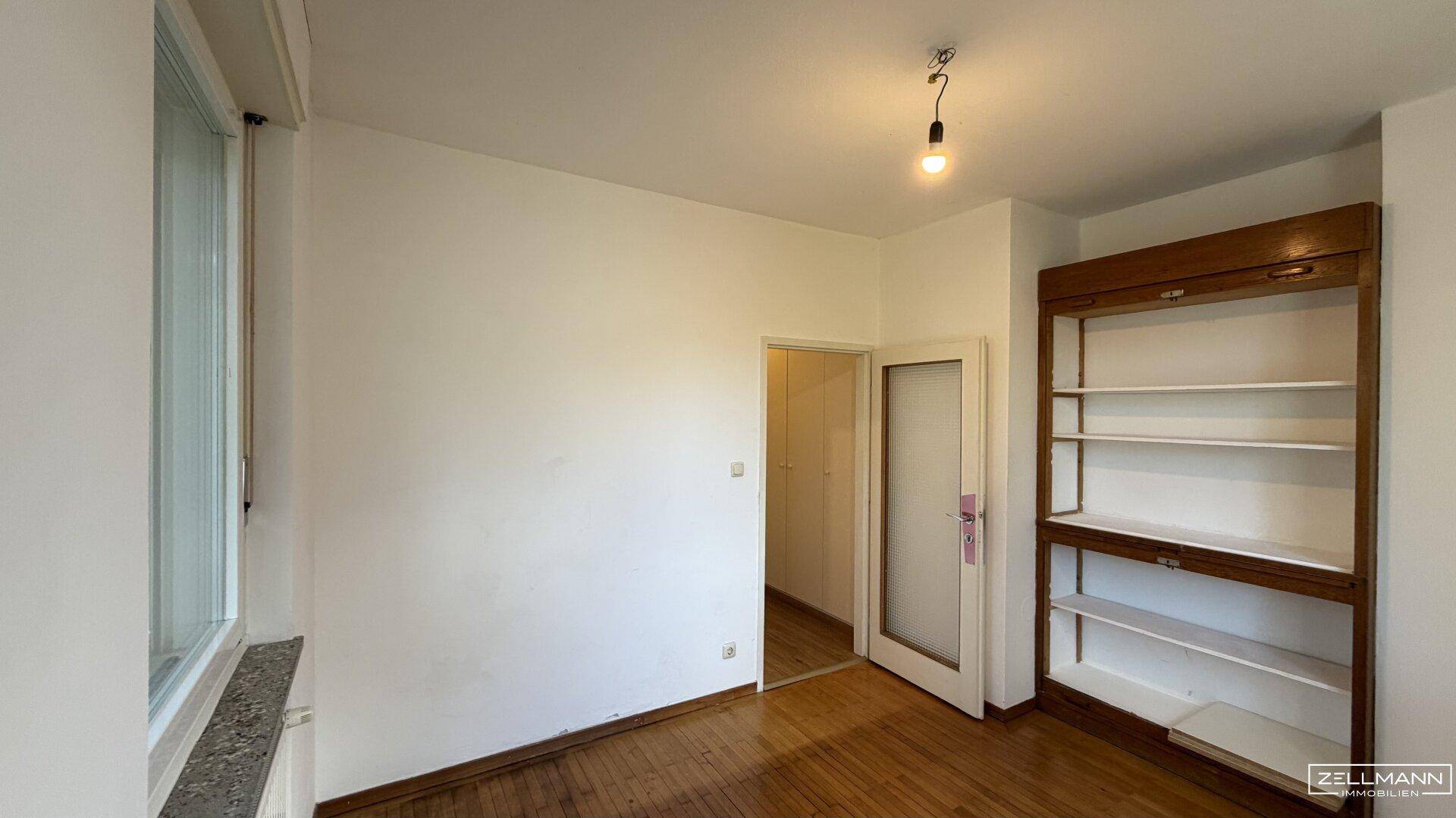 Zimmer mit Parkettboden, Fenster und einem praktischen Einbauregal für Stauraum.