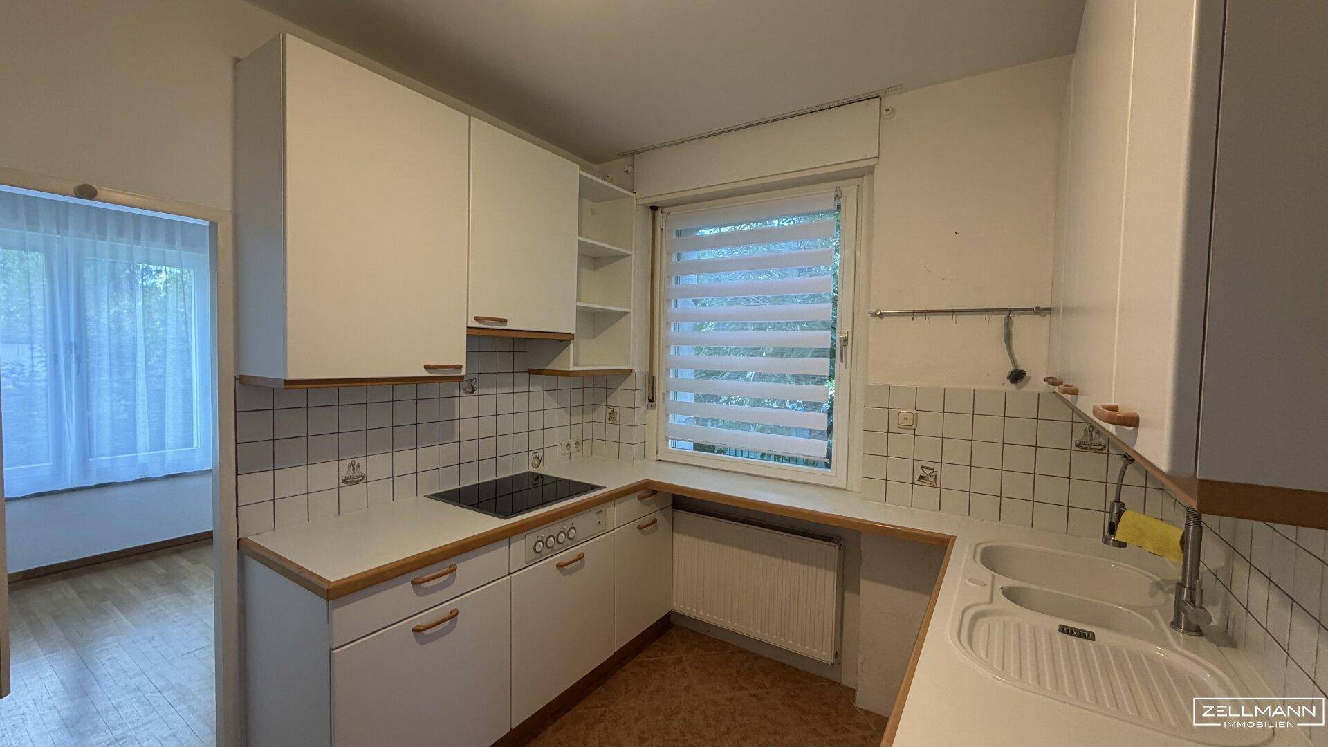 Funktionale Küche mit weißen Schränken, Kochfeld, Spüle und Fenster mit Jalousie.