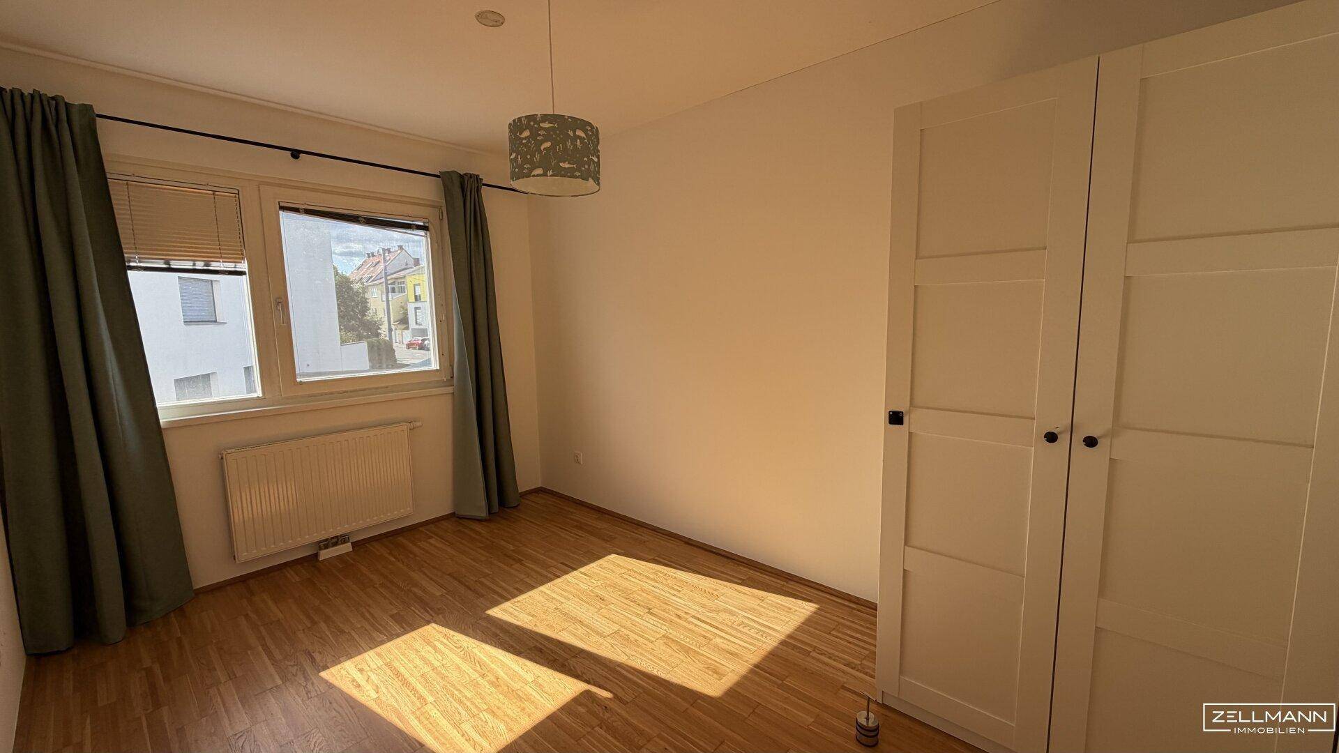 Schlafzimmer mit hellem Holzboden, Fenster und weißem Kleiderschrank, durchflutet von natürlichem Licht.