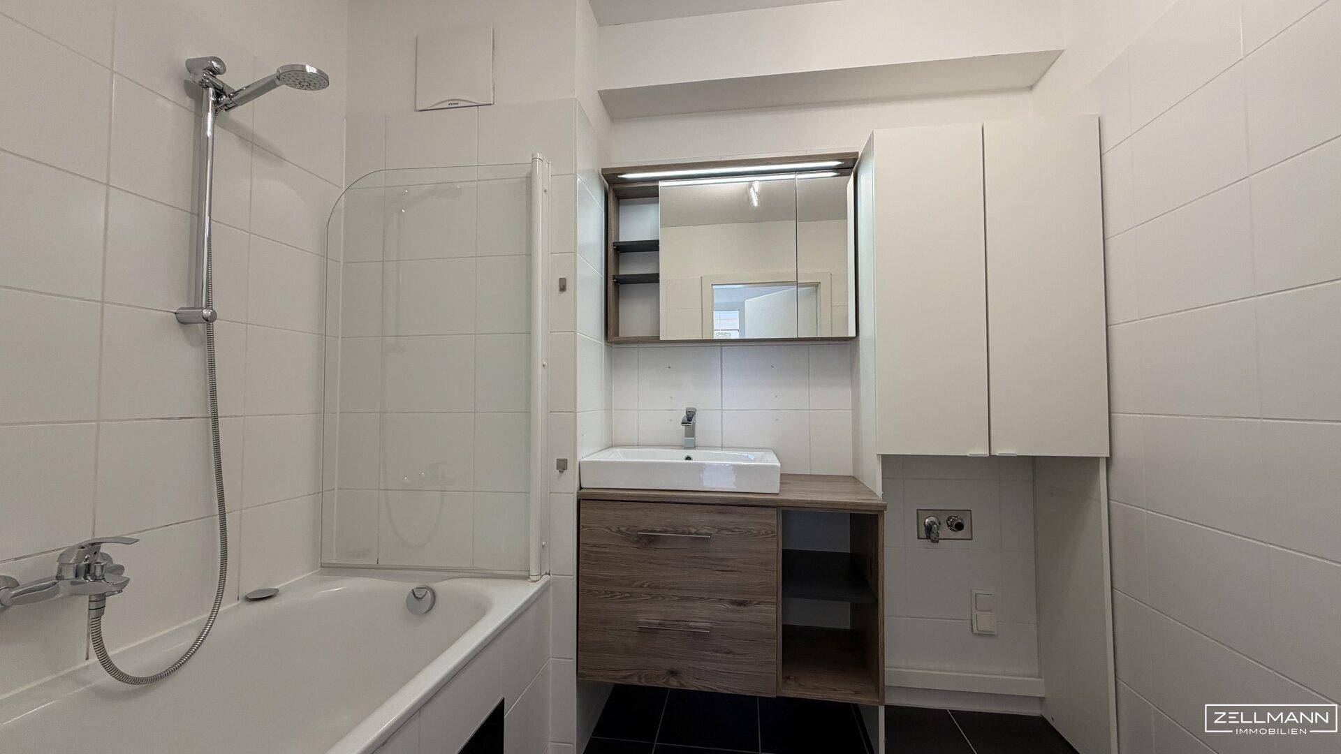 Helles Badezimmer mit Badewanne, Waschtischunterschrank und Spiegelschrank, komplett gefliest in Weiß.