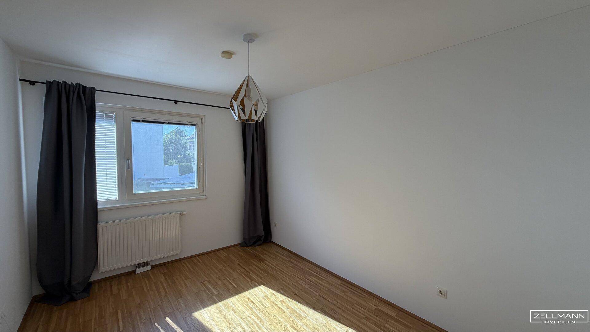 Helles Schlafzimmer mit Holzboden, Fenster und dunklen Vorhängen, ausgestattet mit einer modernen Leuchte.