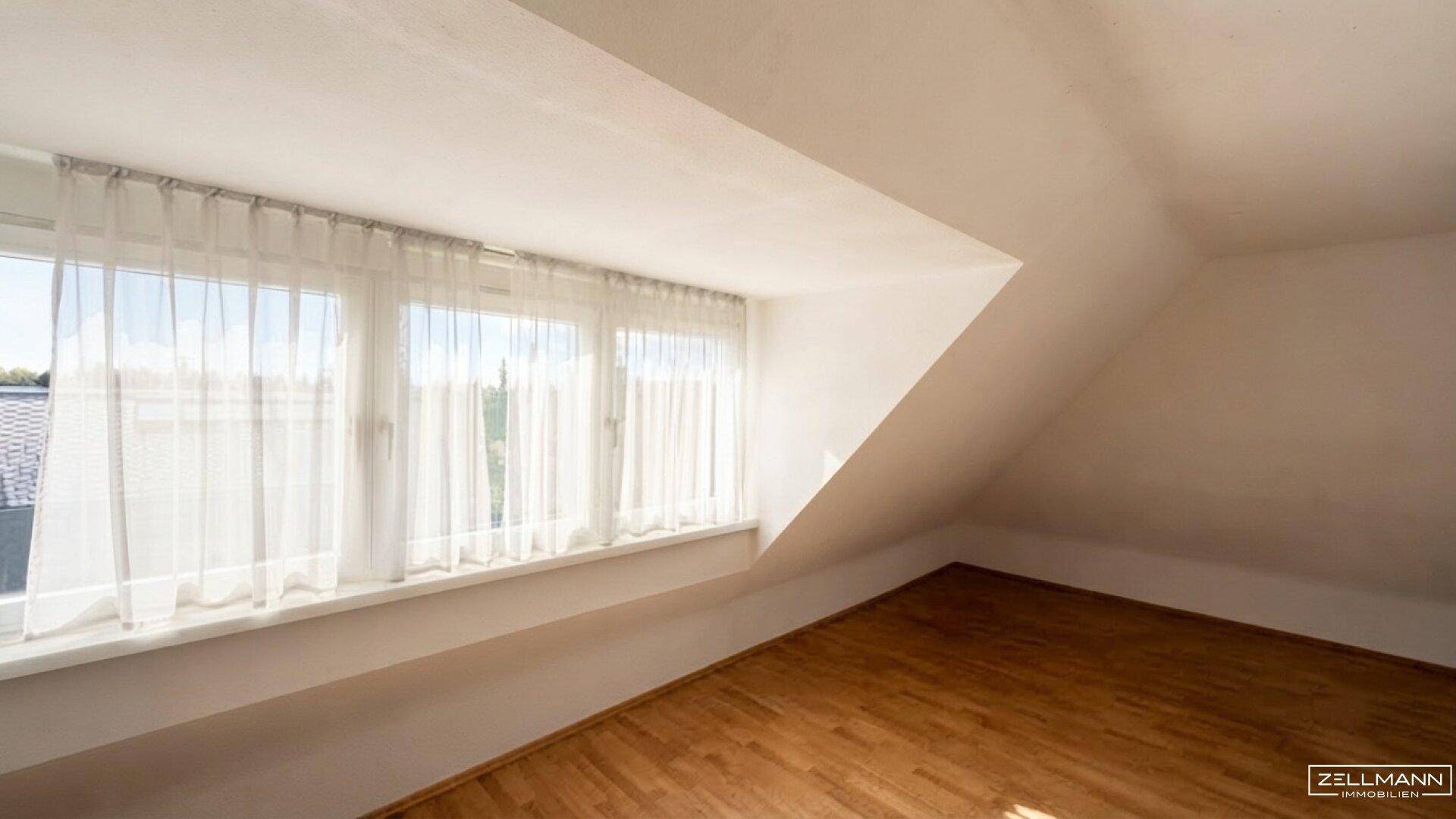 Helles Dachzimmer mit Holzboden und breiter Fensterfront, die viel Tageslicht bietet.