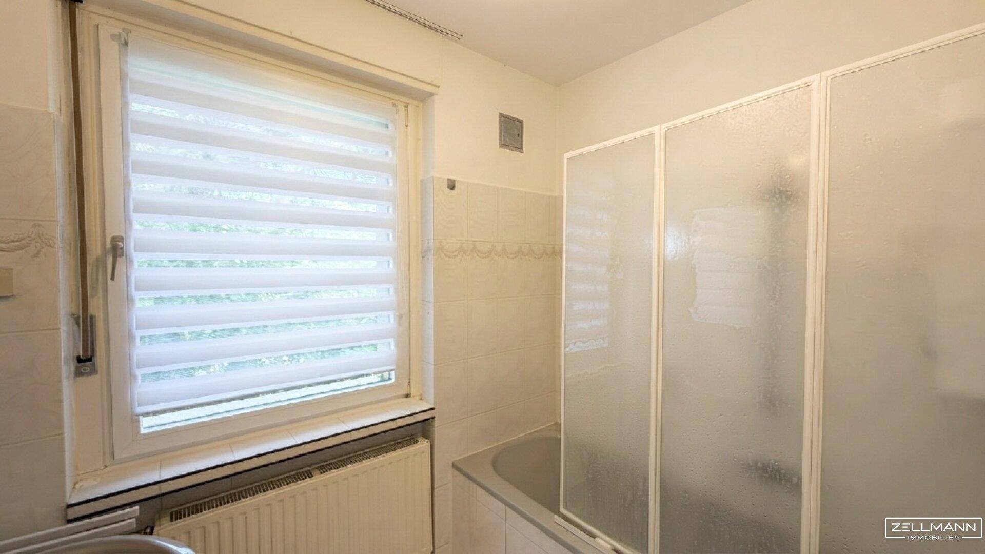 Badezimmer mit Badewanne und Duschabtrennung, Fenster und Fliesen an den Wänden.